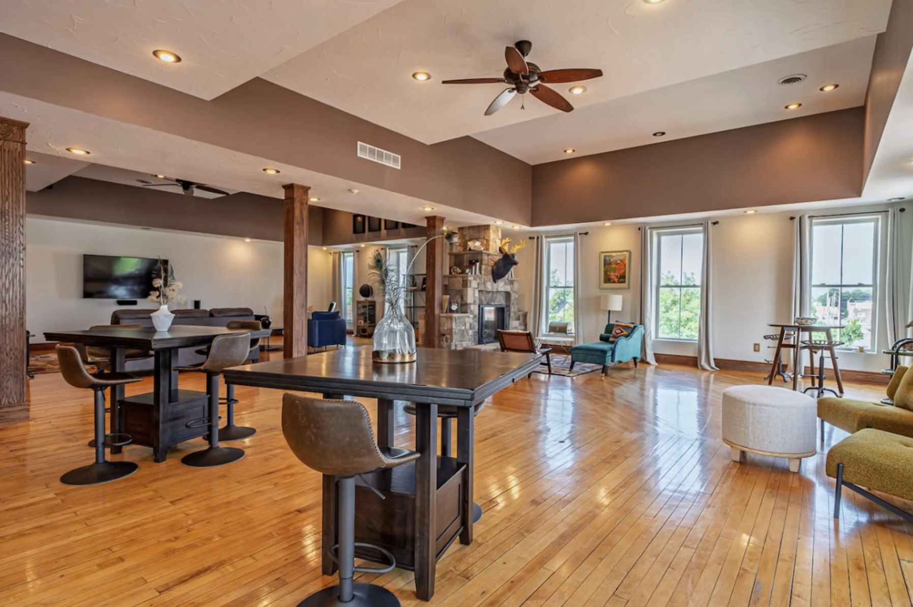 Spacious open-concept living room with hardwood floors, large windows, a stone fireplace, and various seating areas including chairs, a sofa, and a lounge chair, decorated with a mounted deer head and framed artworks, ceiling fan, and multiple ceilin