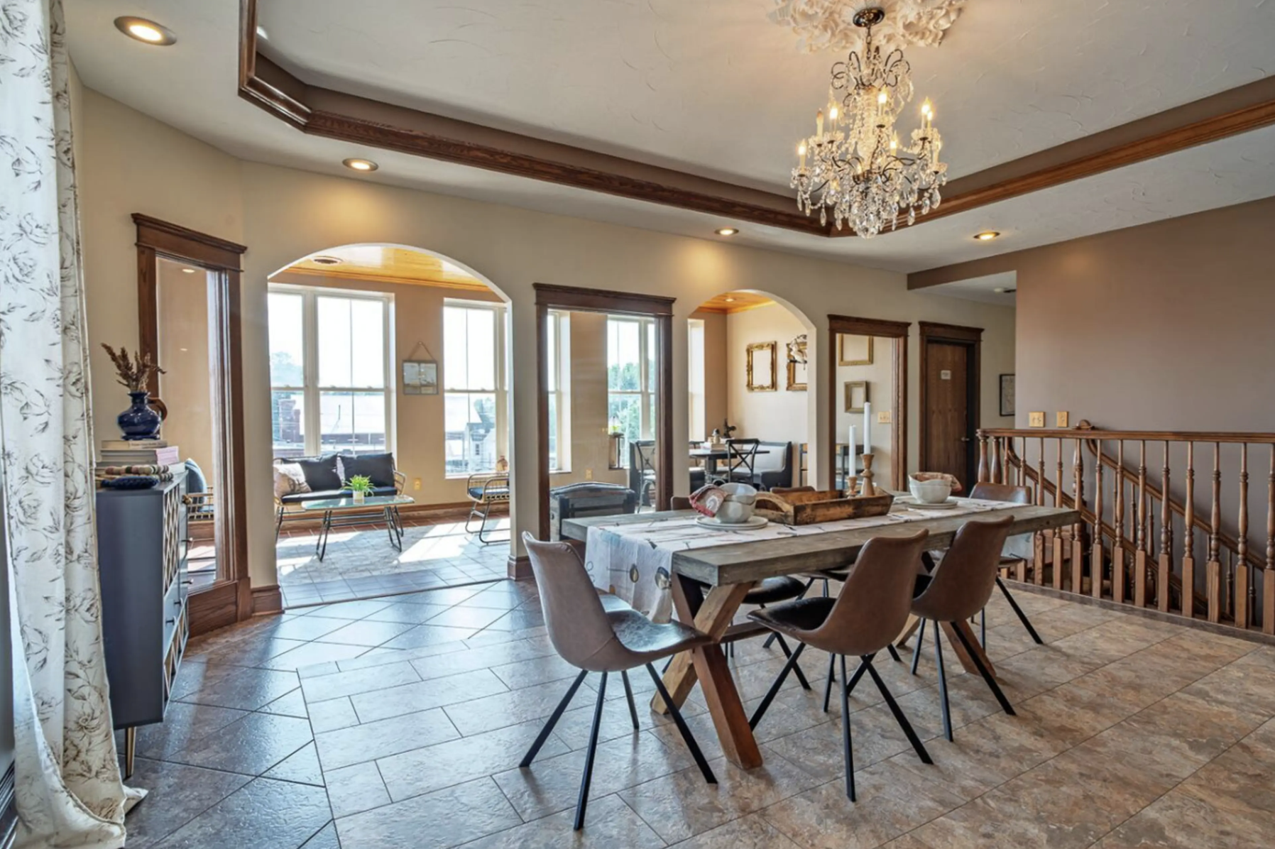 A spacious, well-lit dining area with a rustic wooden table and modern pink chairs. The room features large windows, a chandelier, and decorative wooden trim on the ceiling. There is a staircase with a wooden railing, and a cozy seating nook with a w