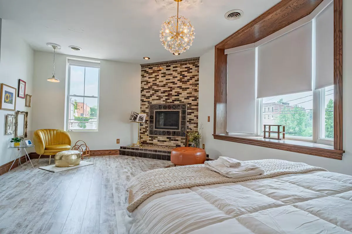 A bright bedroom with a large bed, window with white blinds, fireplace with a brick design, yellow chair, gold ottoman, and modern chandelier