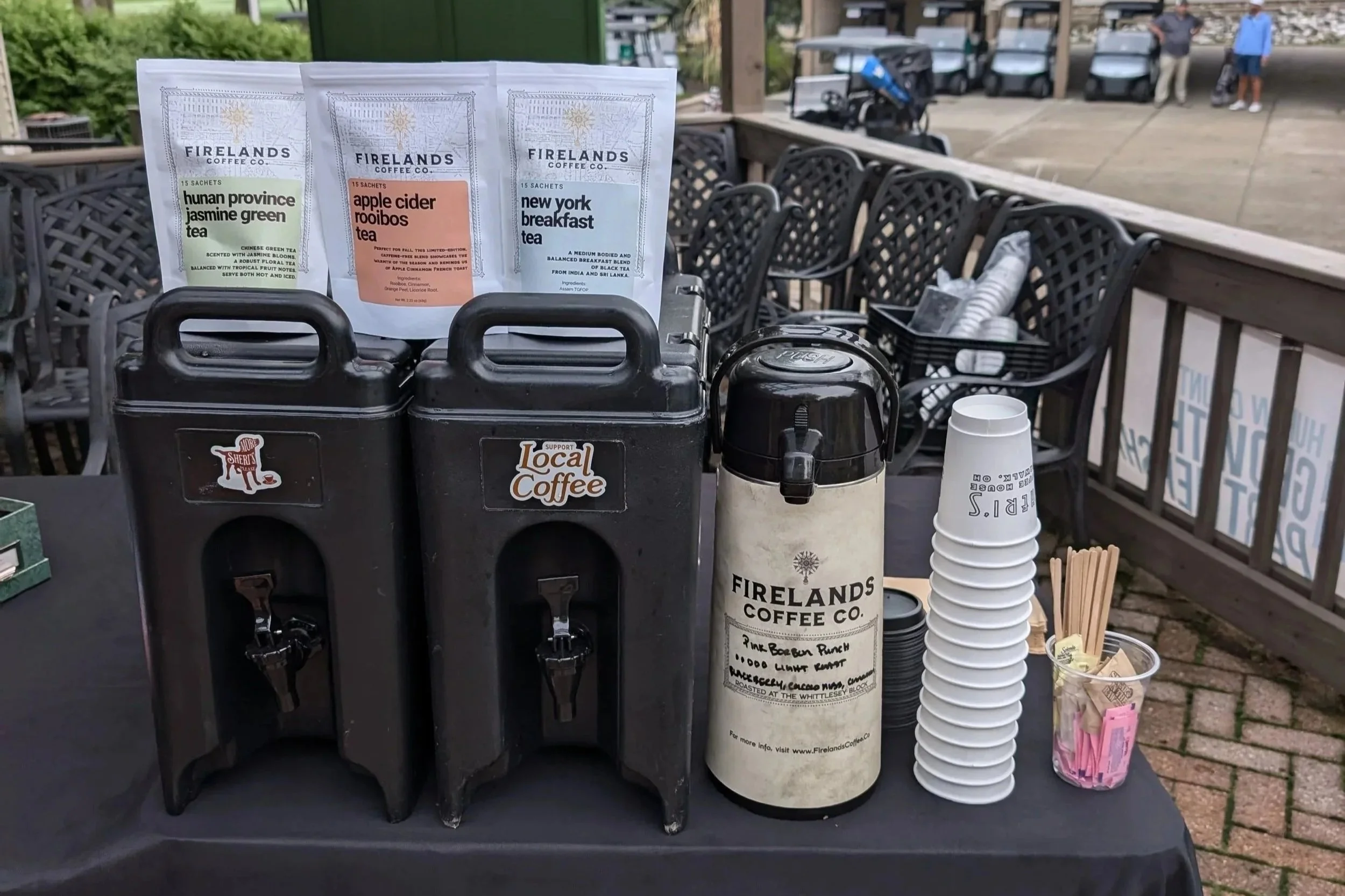 Coffee station with three bags of Firelands Coffee Co. tea varieties, including Hunan Province Jasmine Green Tea, Apple Cider Rooibos Tea, and New York Breakfast Tea, along with a hot water dispenser, disposable cups, and stir sticks on a black table outdoors.