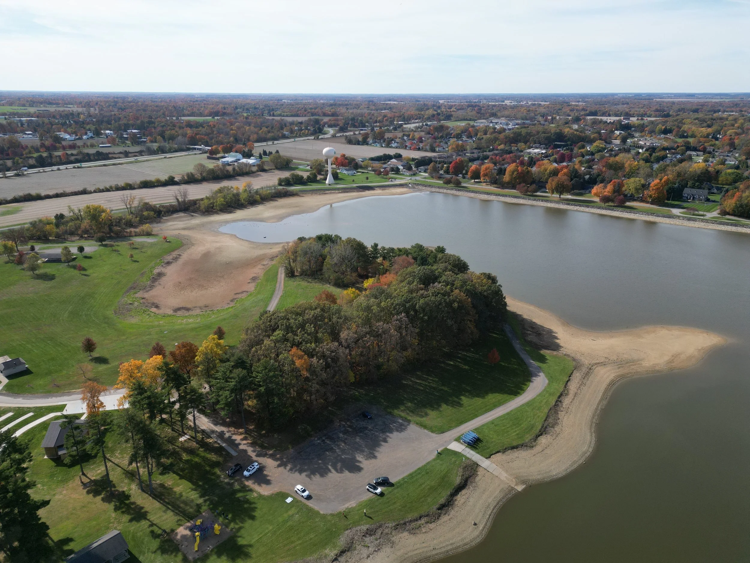 Aerial view of a park with a lake, playground, and parking lot, surrounded by trees with autumn foliage, and residential area in the background.
