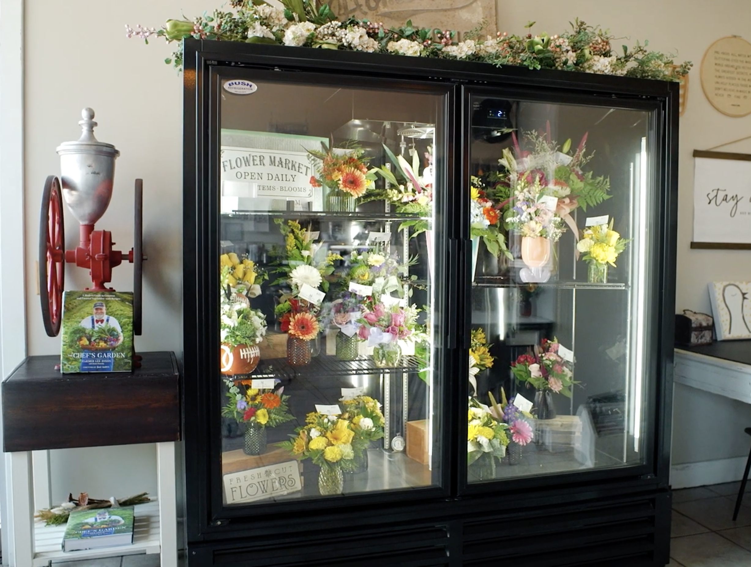 A display case filled with various colorful flower bouquets inside a floral shop. A sign reading 'Fresh Cut Flowers' is visible inside the case. On top of the case, there are more floral arrangements. To the left, a vintage red and silver fire alarm is mounted on the wall next to a brochure stand with a gardening book. The background includes part of a wall with a wooden plaque and a framed quote.