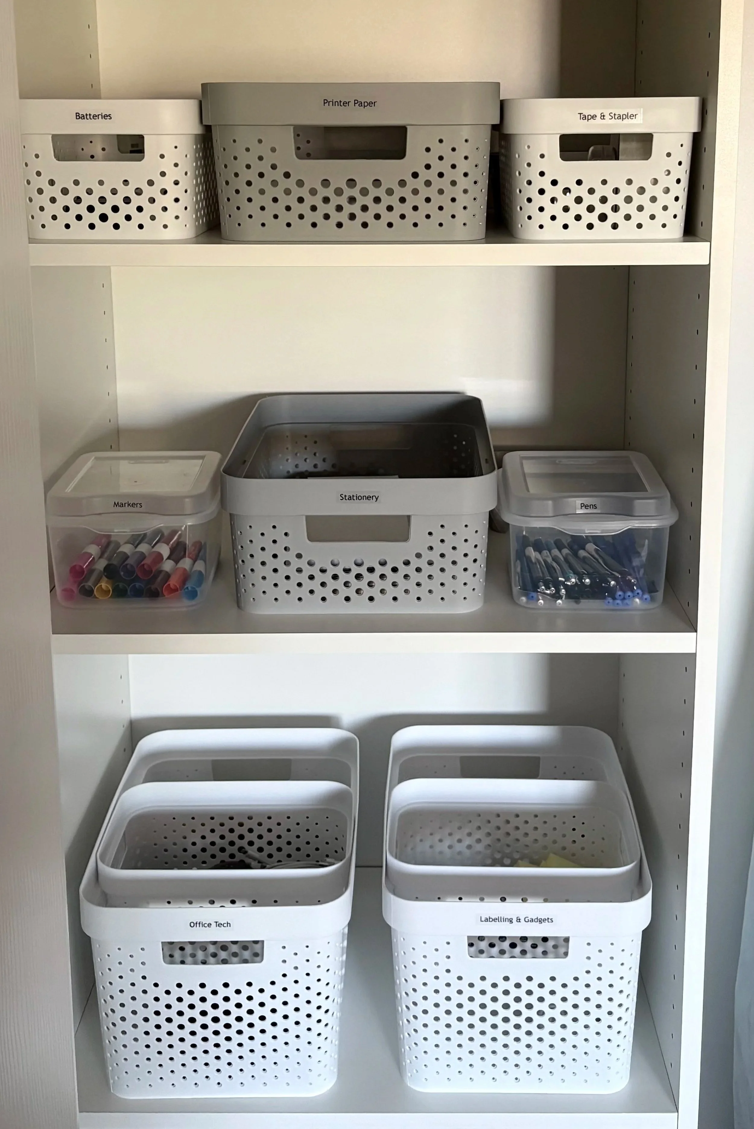 A shelving unit with labeled opaque and clear storage bins containing office supplies, stationery and technology gadgets.