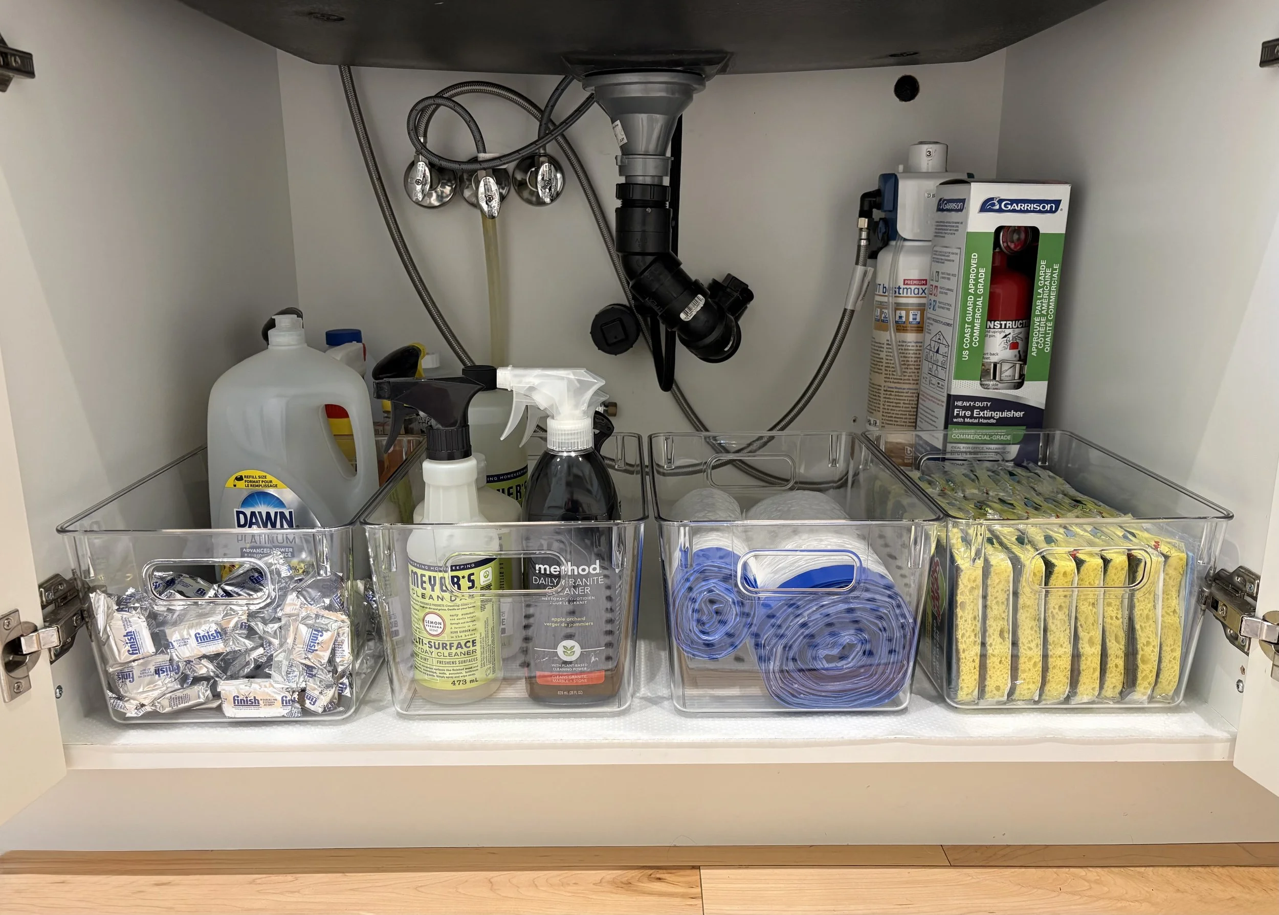 Inside of a tidy kitchen cabinet with cleaning supplies and kitchen essentials organized in clear storage bins.
