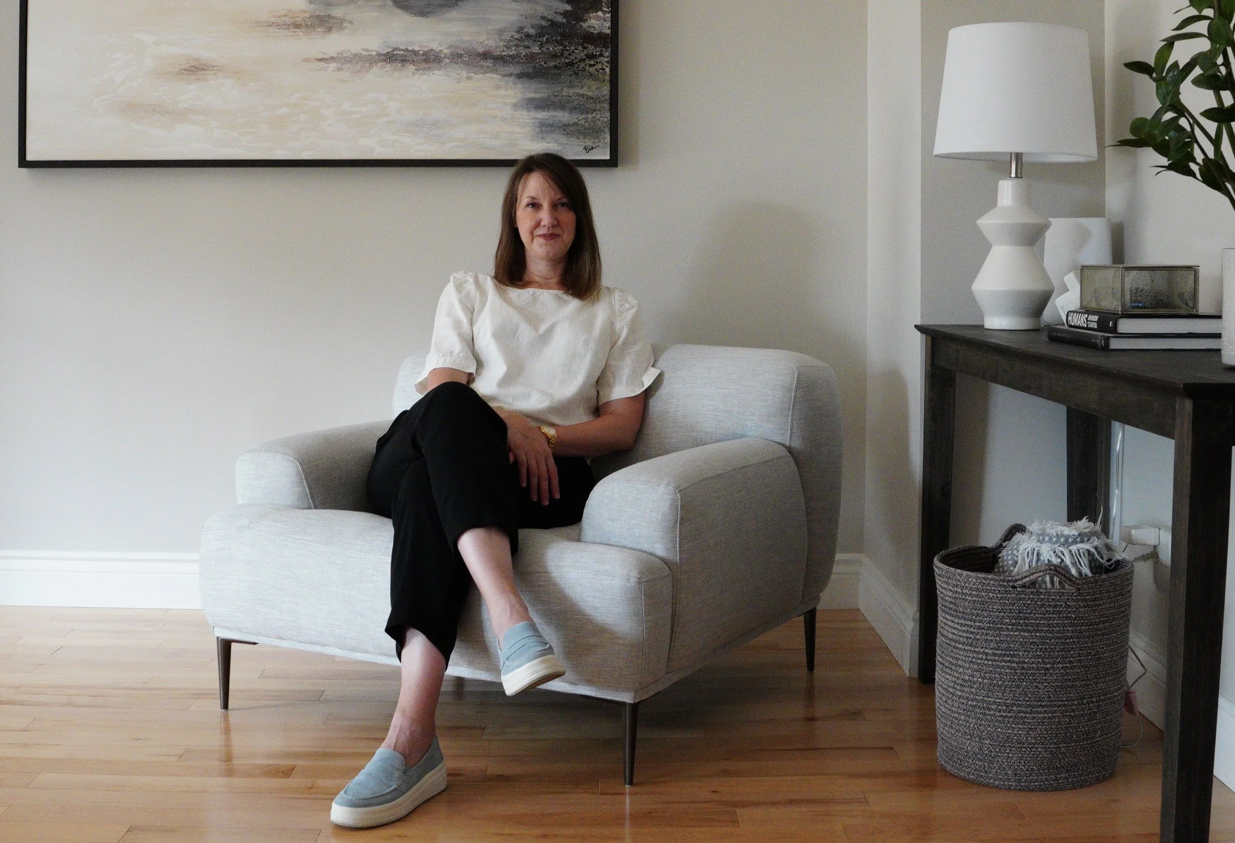 A woman sitting on an arm chair in a living room. Behind her is a console table with a plant and books neatly arranged.