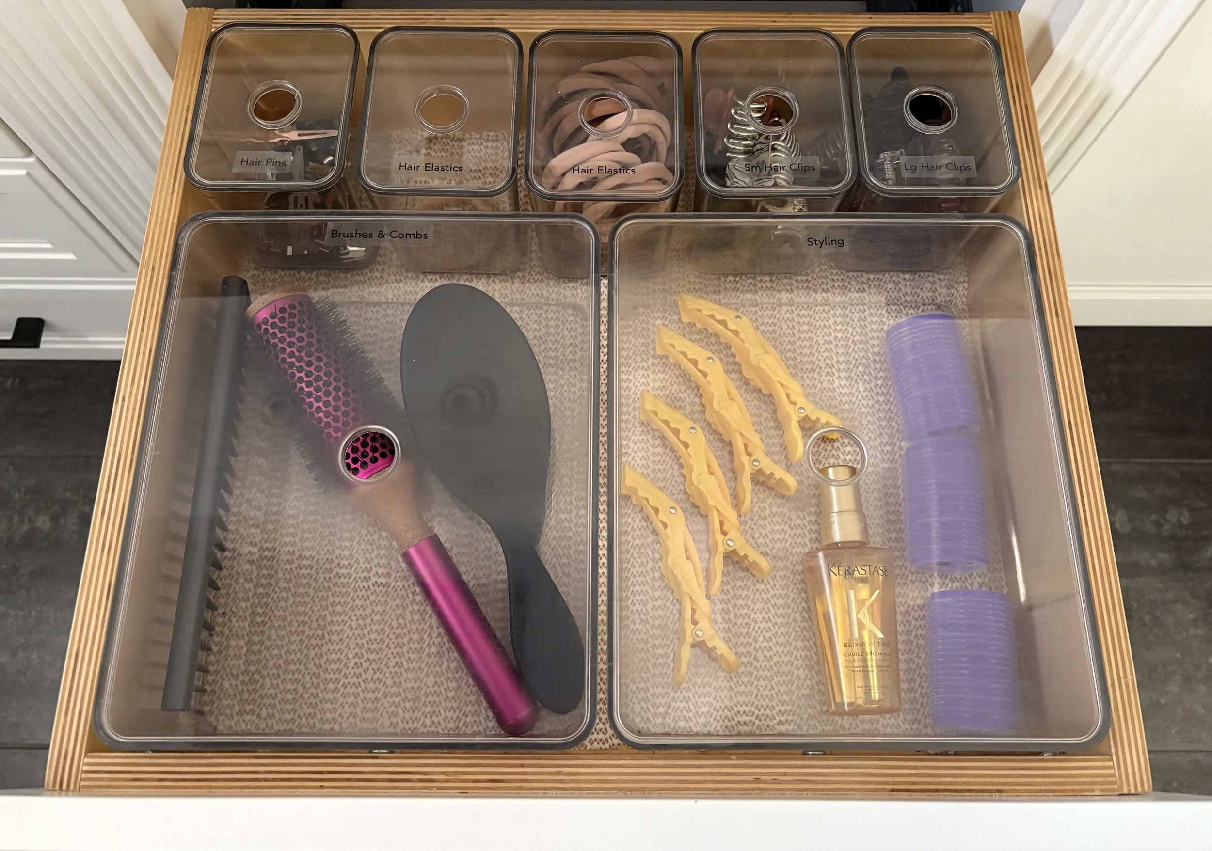 An open bathroom vanity drawer with assorted sizes of labeled clear plastic containers holding hair styling tools.