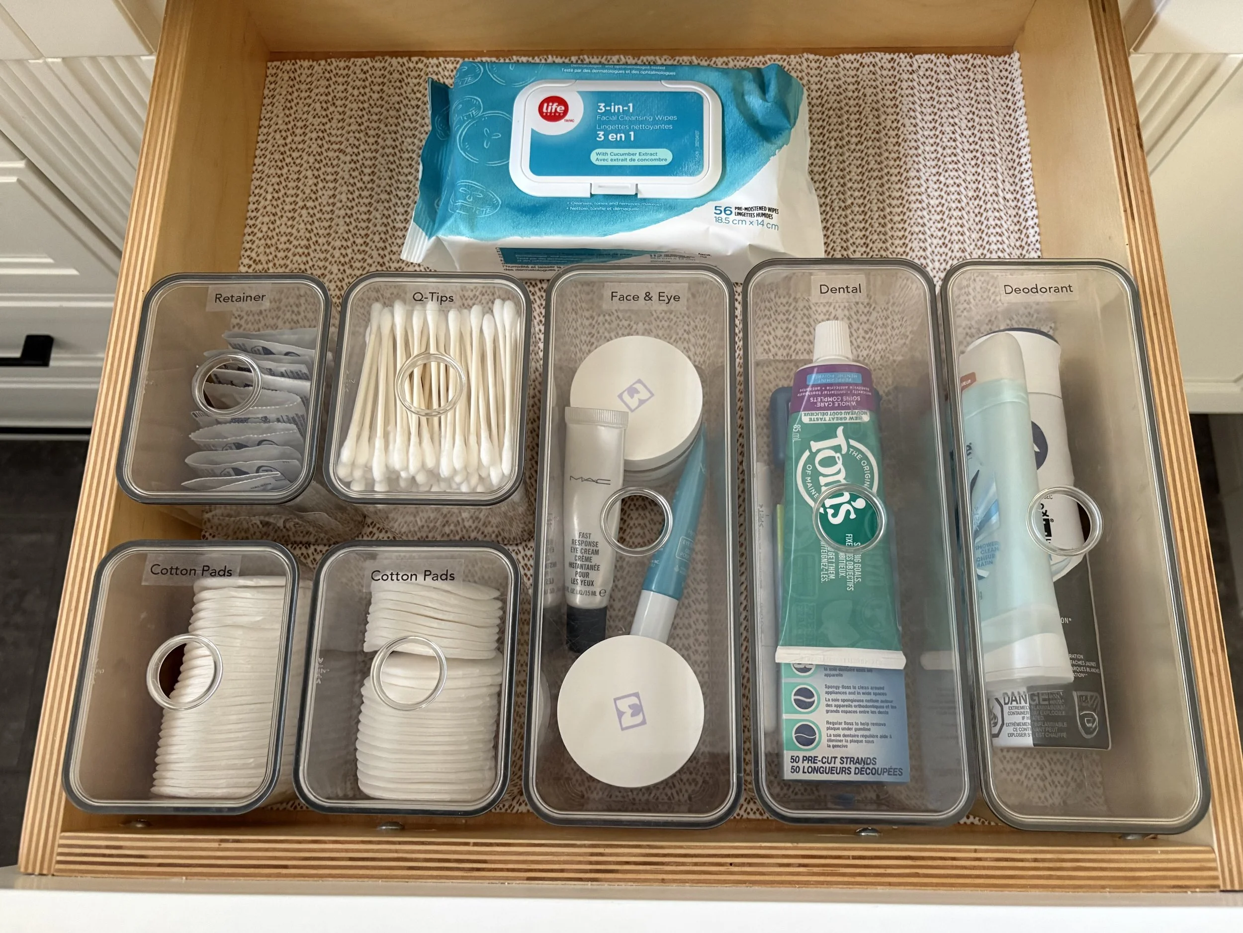 An open bathroom vanity drawer with assorted sizes of labelled clear plastic containers holding personal care products