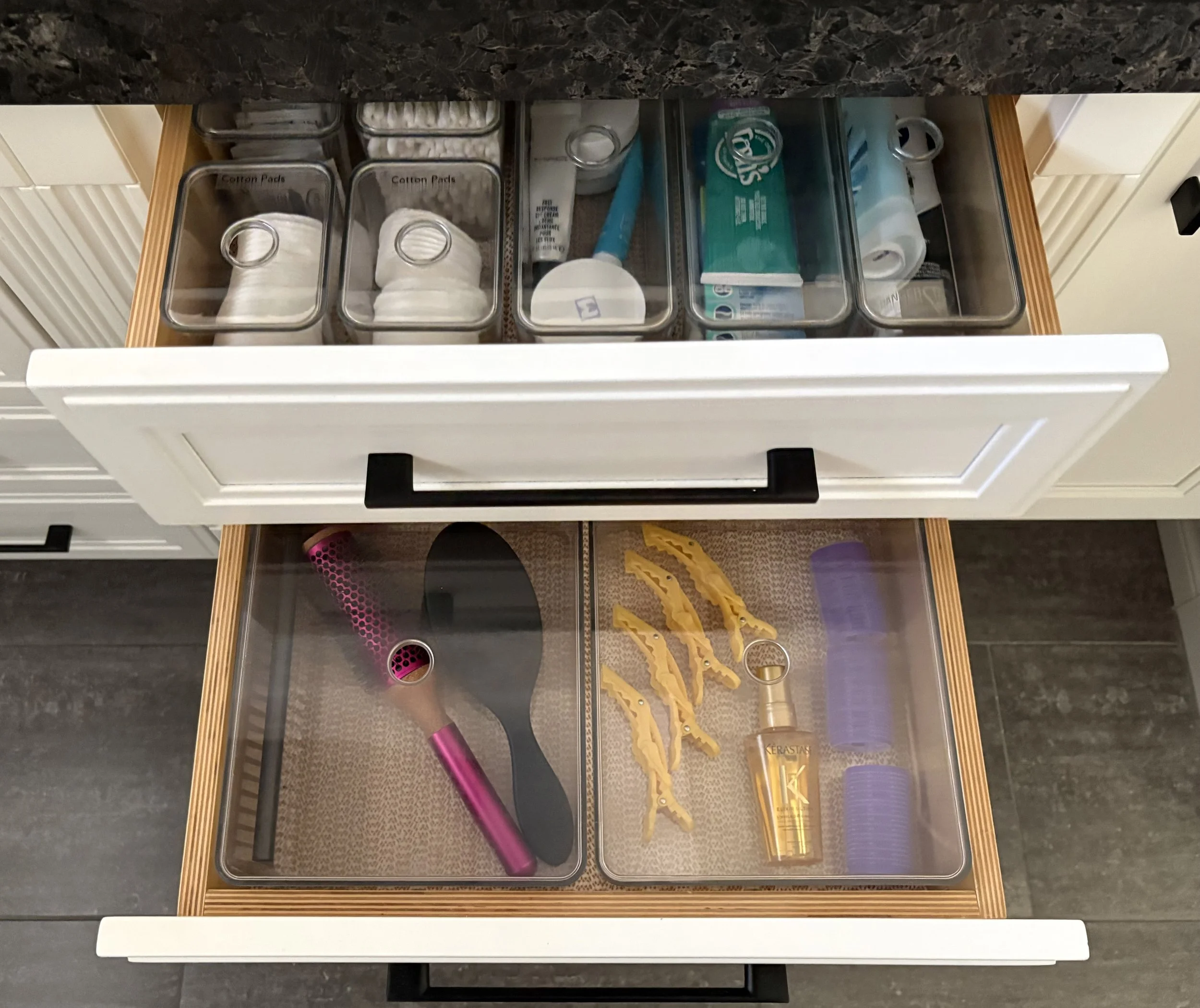 Two open bathroom vanity drawers with labelled clear plastic containers holding personal care and hair styling products.