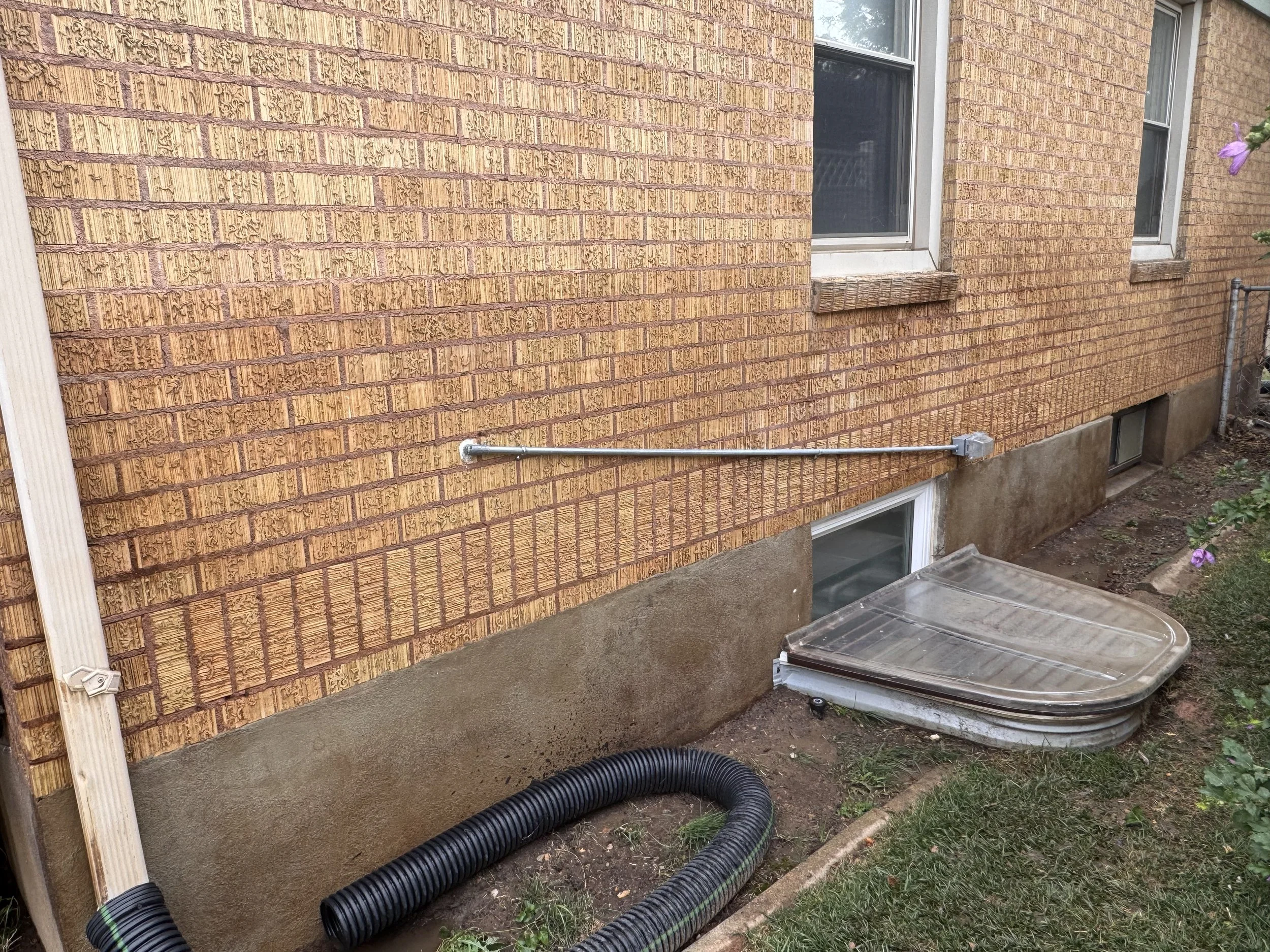 The image shows the exterior of a brick house with two windows. There is a black corrugated drainage pipe on the ground, and part of a wooden post and a garden hose are also visible. The house has a concrete foundation and some garden plants are grow