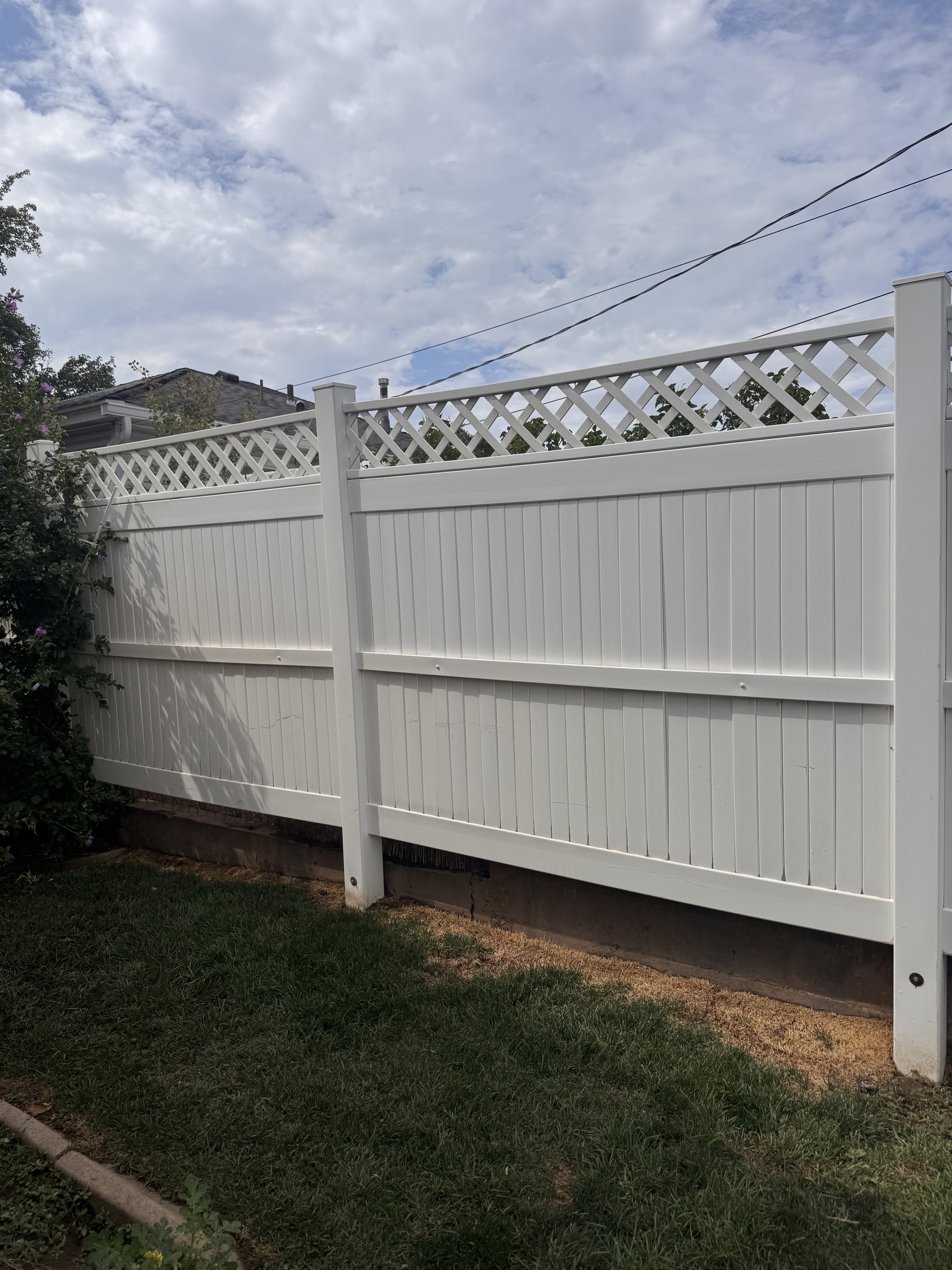 A white privacy fence with lattice top, partially hiding the house behind it, next to a grassy yard and some garden bushes, under a partly cloudy sky.