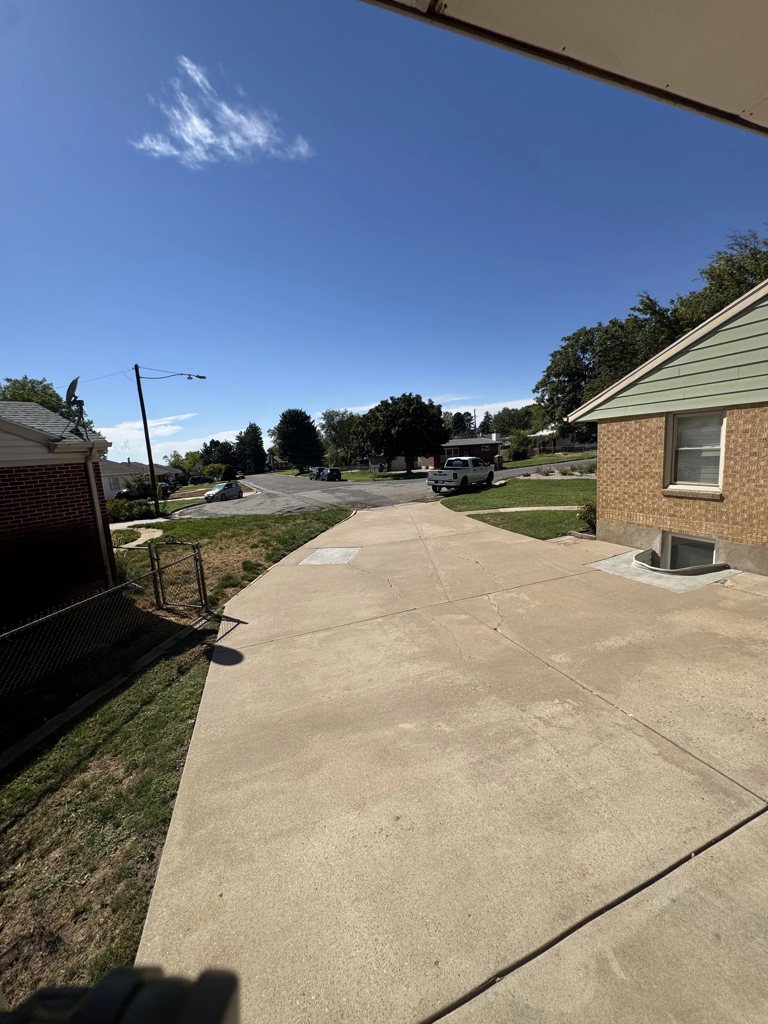 View of a suburban neighborhood street with a concrete sidewalk, grass patches, houses, and parked cars under a blue sky with a few clouds.