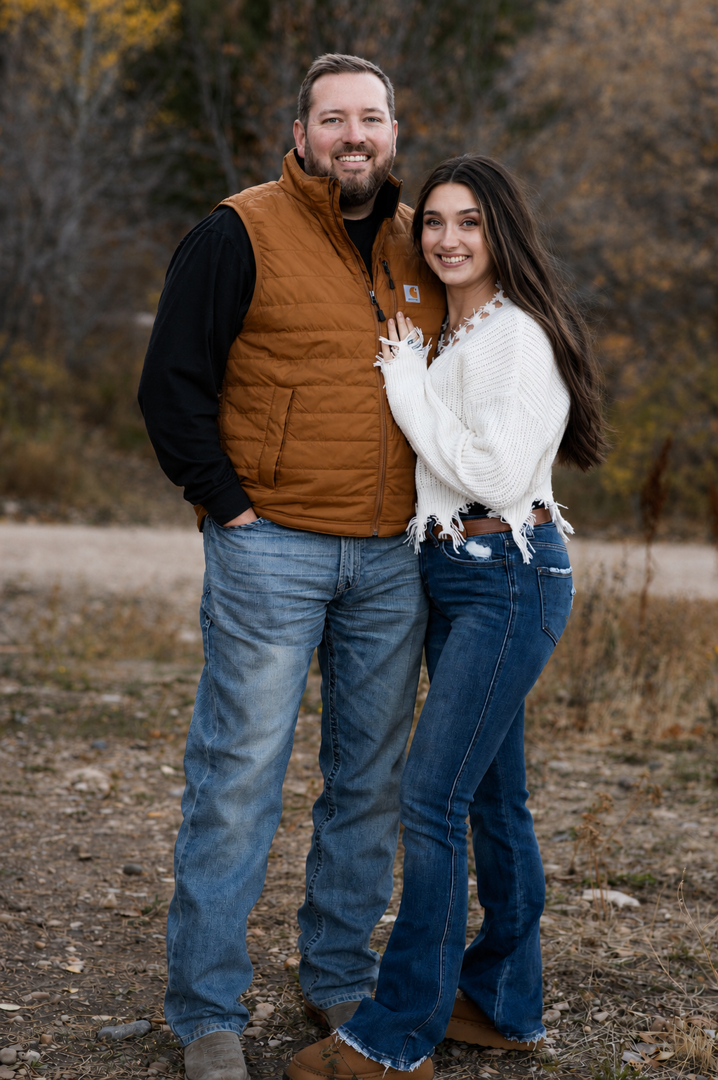 A smiling couple stands outdoors in an autumnal setting, with trees and fallen leaves in the background. The man is wearing a brown puffer vest over a black long-sleeve shirt and jeans. The woman is wearing a white, distressed sweater and blue jeans.