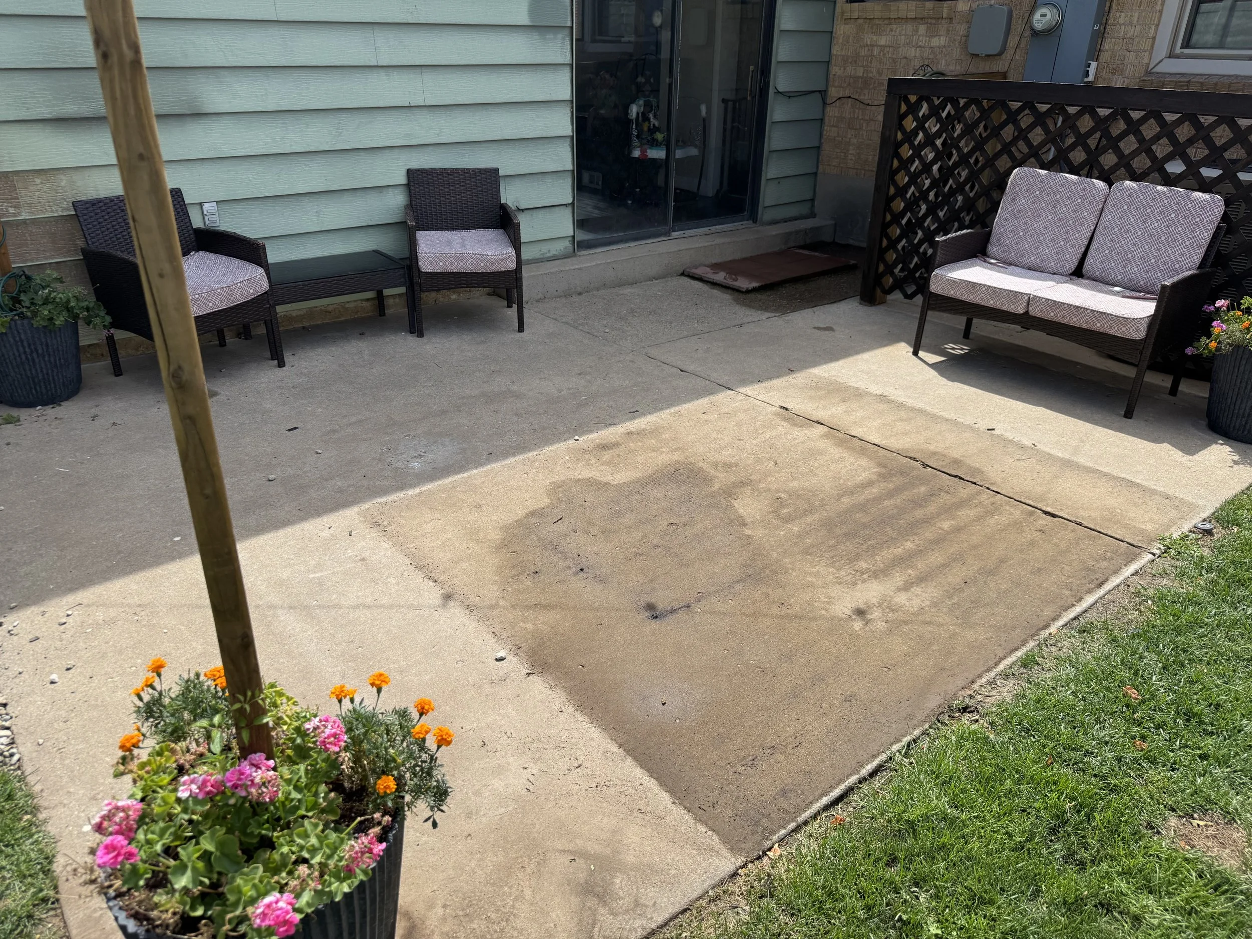 A backyard patio with a flower pot filled with pink and orange flowers on the left, a small sitting area with two chairs, a side table, and a loveseat, with an sliding glass door leading to the house, and a stone pathway leading away from the patio.