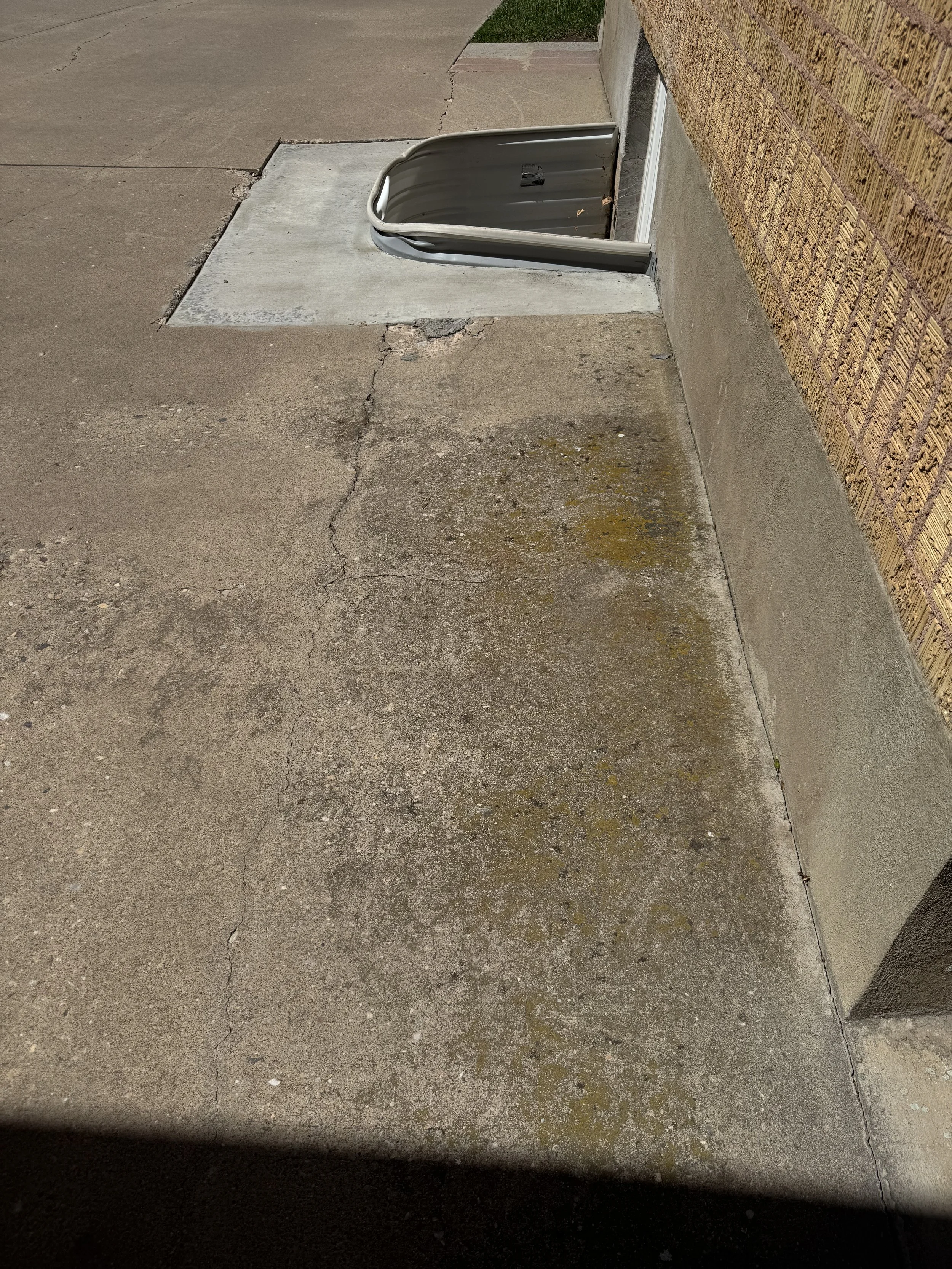 An outdoor scene showing a cracked concrete sidewalk with a partially open metal vent or drain on the ground next to a brick wall, with some dirt and moss around the vent.