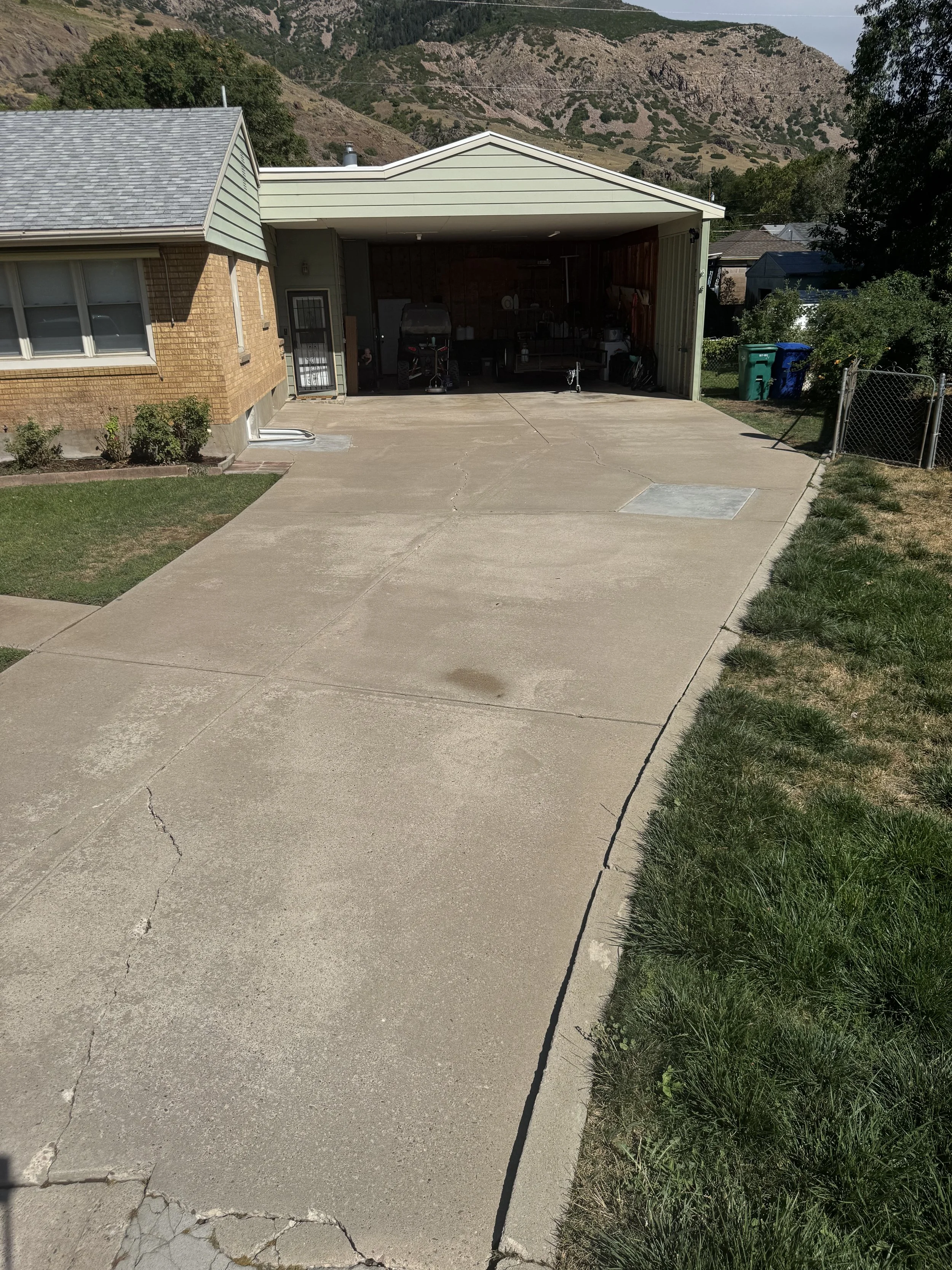 A residential driveway with a garage behind, mom and child in the garage, and mountains in the background. The driveway is cracked and partially shaded, with a small garden on the left and grass on the right.