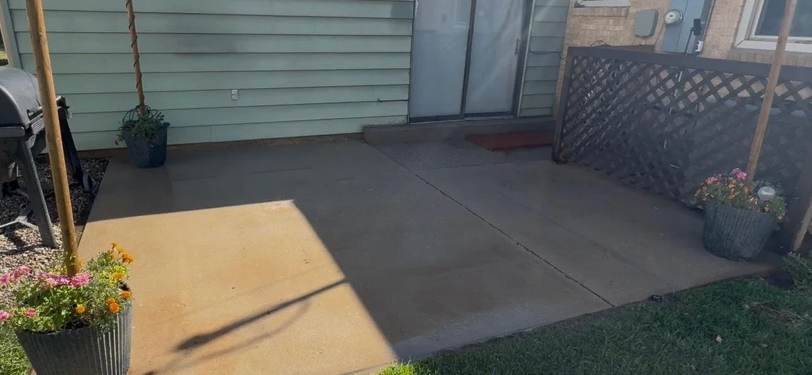 A small backyard patio with concrete floor, three flower pots with colorful flowers, a black grill, and a lattice fence with a potted plant, adjacent to a house with light blue siding and a sliding glass door.