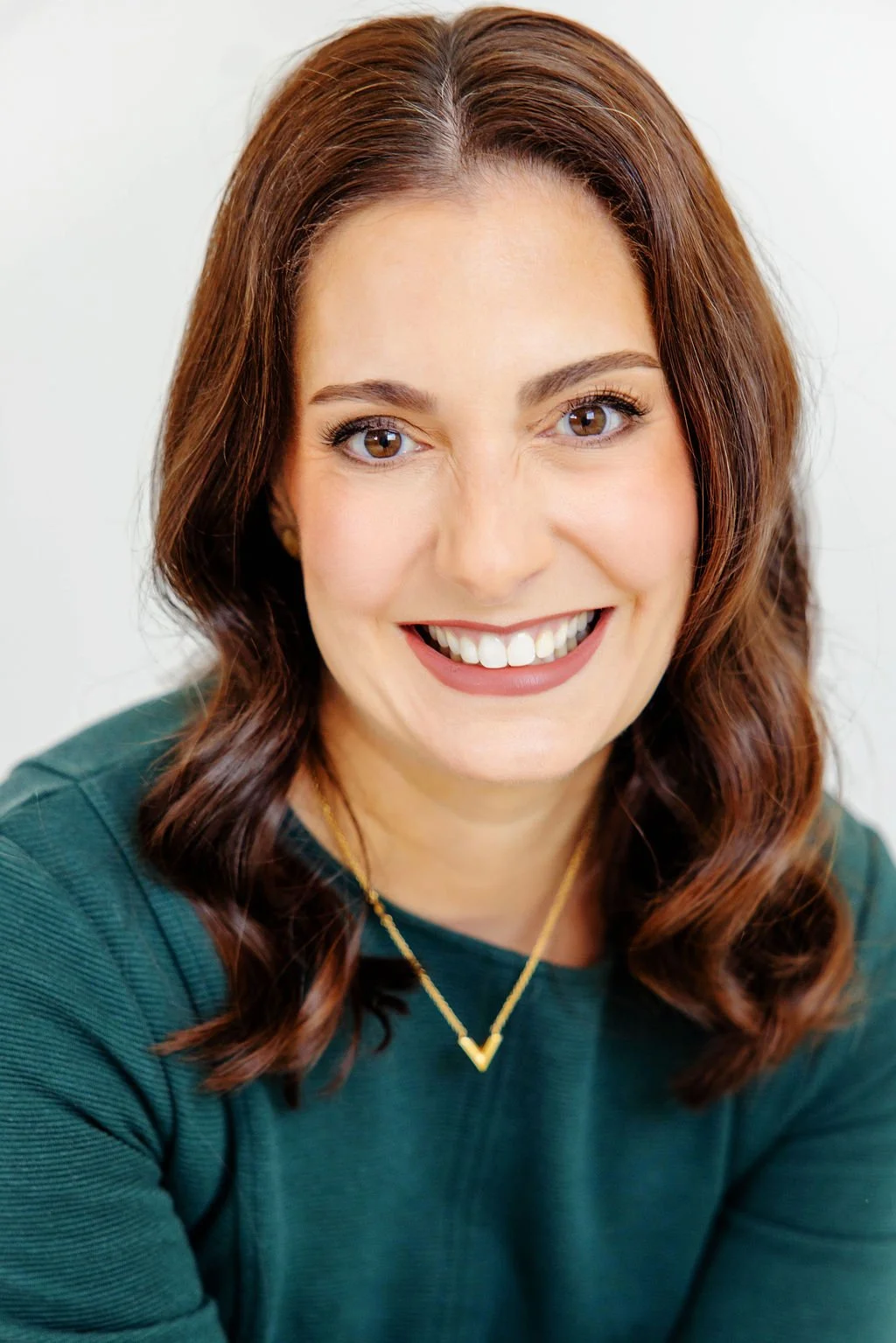 Close-up headshot of Alison De Villiers smiling with brown hair and brown eyes, wearing a green top and a gold necklace.