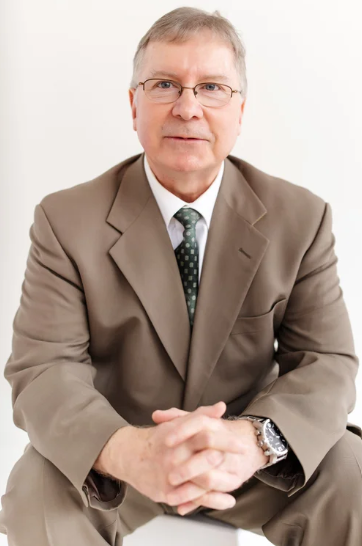 J Scott Mullins smiling, wearing a beige suit, white shirt, and dark tie, sitting with his hands clasped, a plain white background.