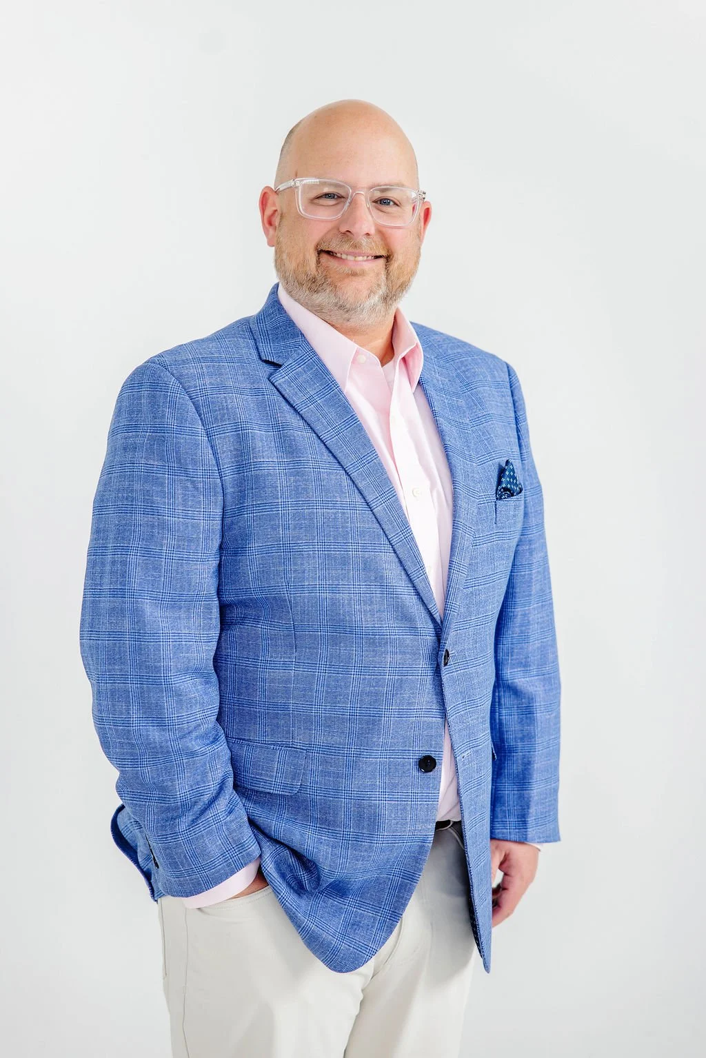 Brian Jones smiling, glasses and a beard, wearing a blue plaid blazer, pink shirt, and beige pants, standing against a plain white background.