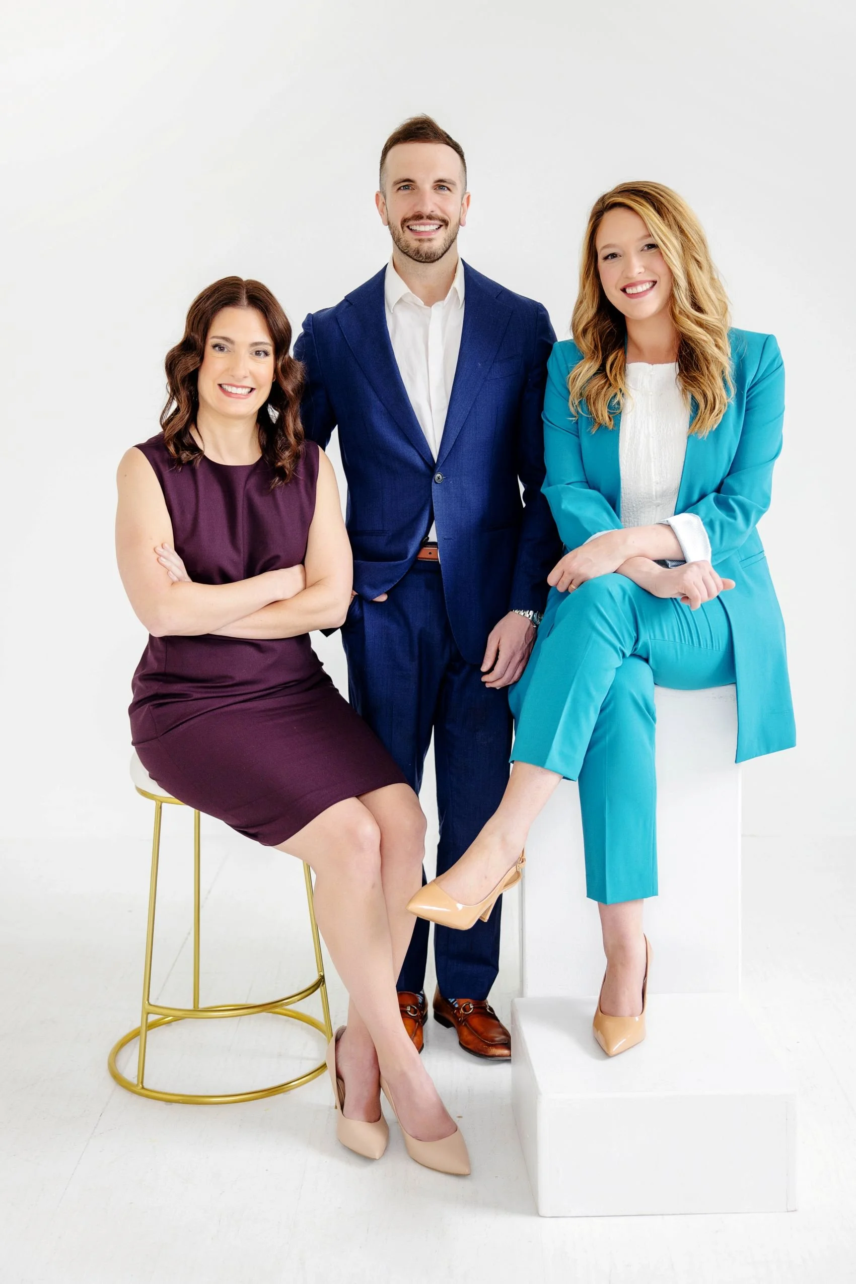 , happy and smiling, dressed in business attire against a white background. One woman is seated on a gold stool, the man and the other woman are standing, with the seated woman crossing her arms and the woman standing on the right sitting with her legs crossed. The man is wearing a blue jacket, and the women are wearing a purple dress and a turquoise pantsuit, respectively.
