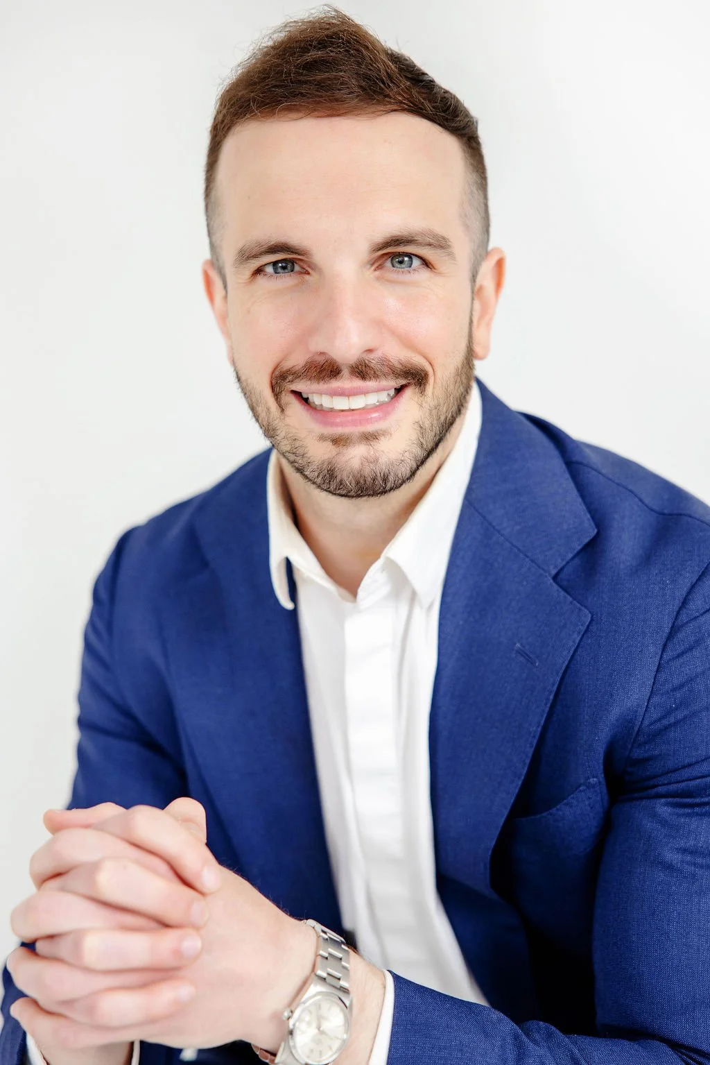 Headshot of Austin LiPuma smiling with short brown hair and a beard, wearing a blue blazer over a white shirt, sitting with hands clasped, against a plain white background.