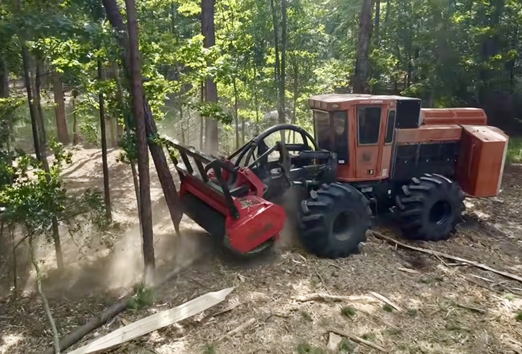 A red forestry or utility tractor with large tires working through a wooded area, moving dirt or debris with a front attachment.