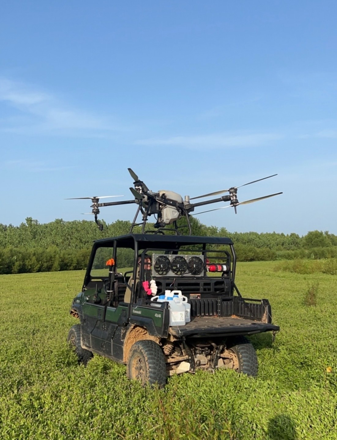 A black utility vehicle with a drone mounted on top, sitting in a grassy field with a background of trees and a clear blue sky.