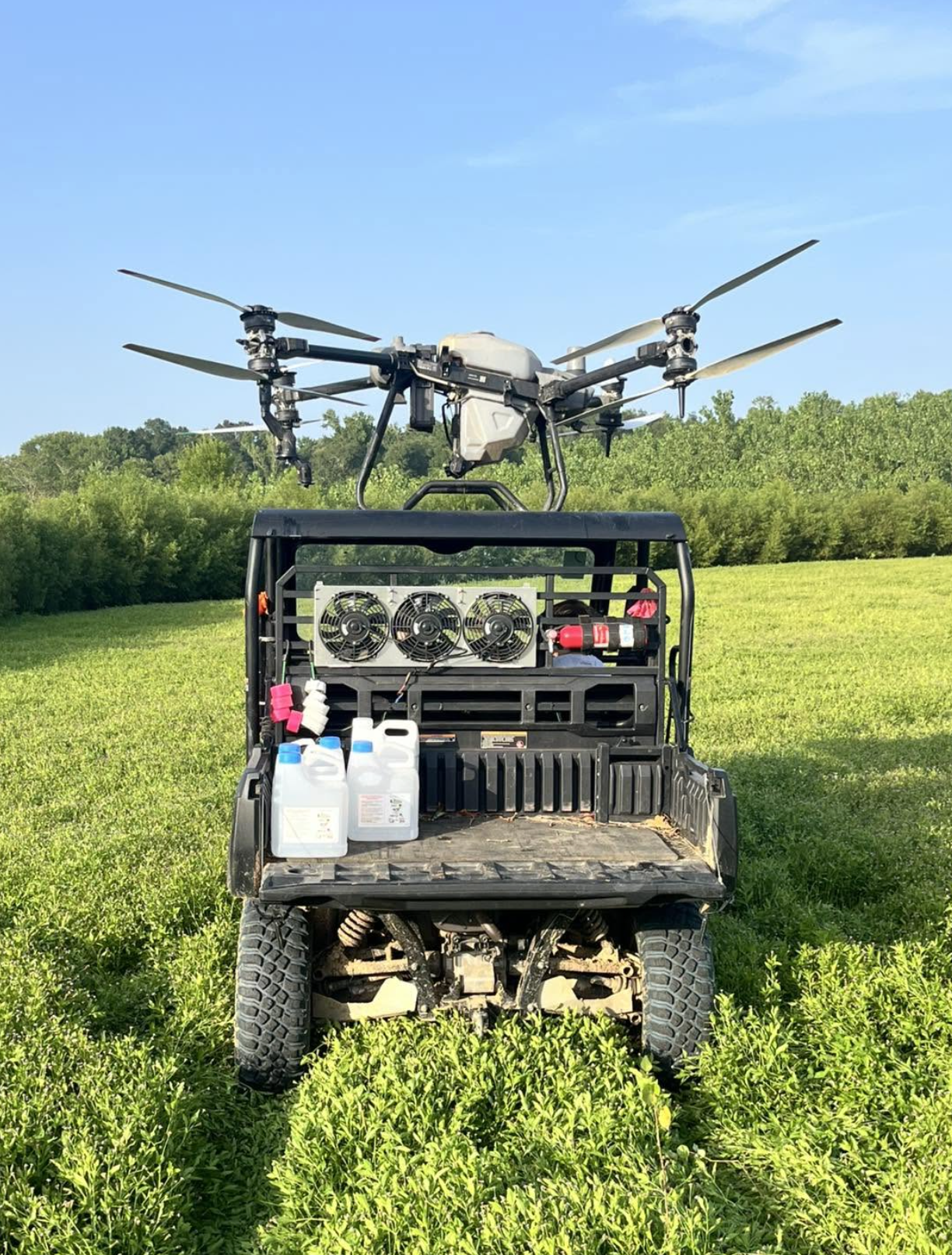 A small off-road utility vehicle with a mounted drone on top, set in a grassy field with green trees under a blue sky.