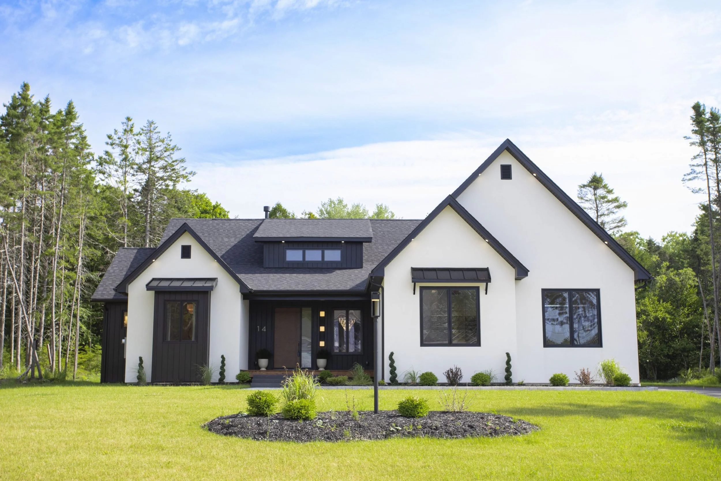 Front view of a modern two-story house with a black roof, white walls, and black window frames, surrounded by a green lawn and trees, under a partly cloudy sky.