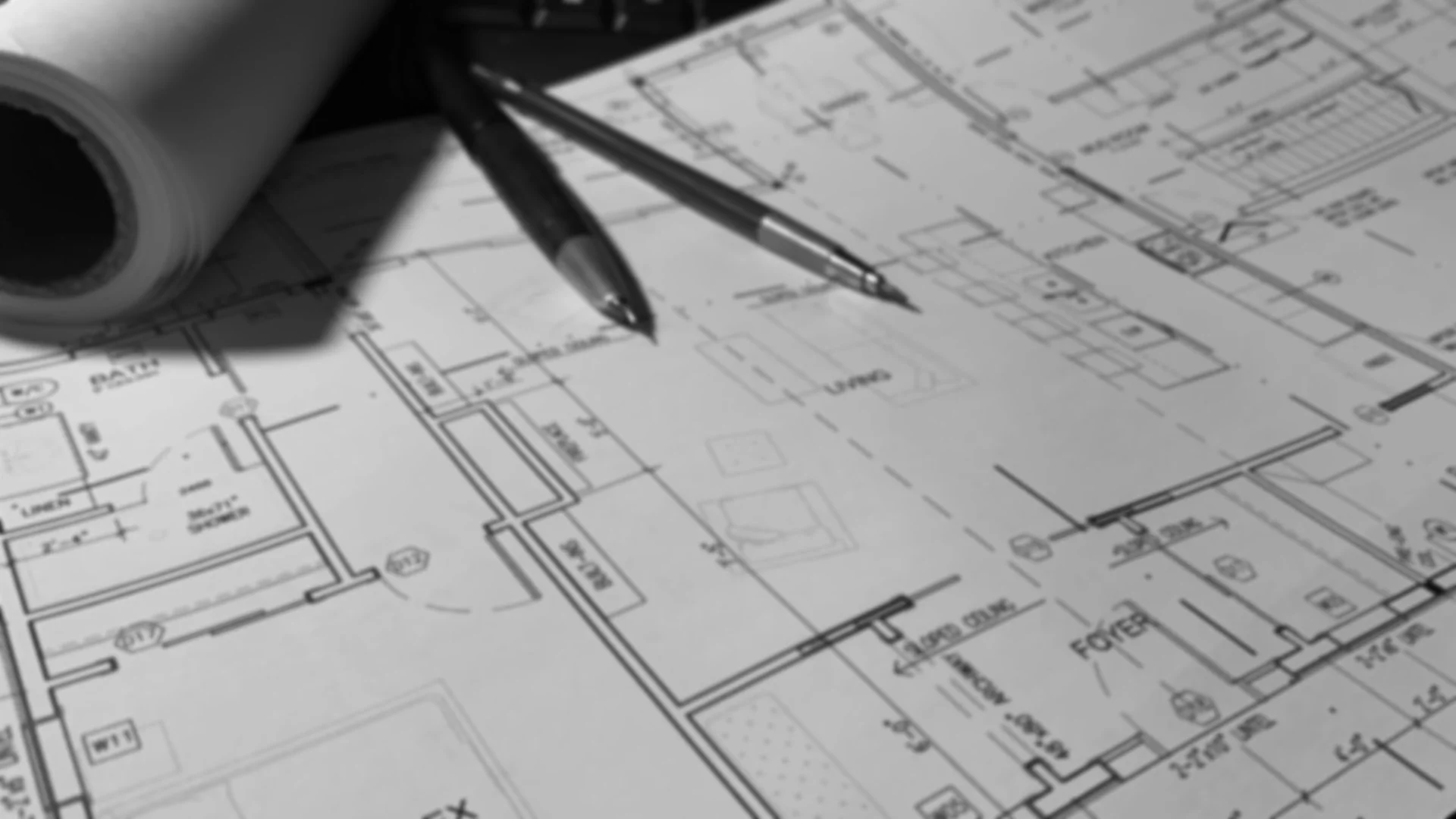 Black and white photo of drafting house plans architectural blueprints on a table with a rolled-up blueprints, a pen, and a drafting pencil.