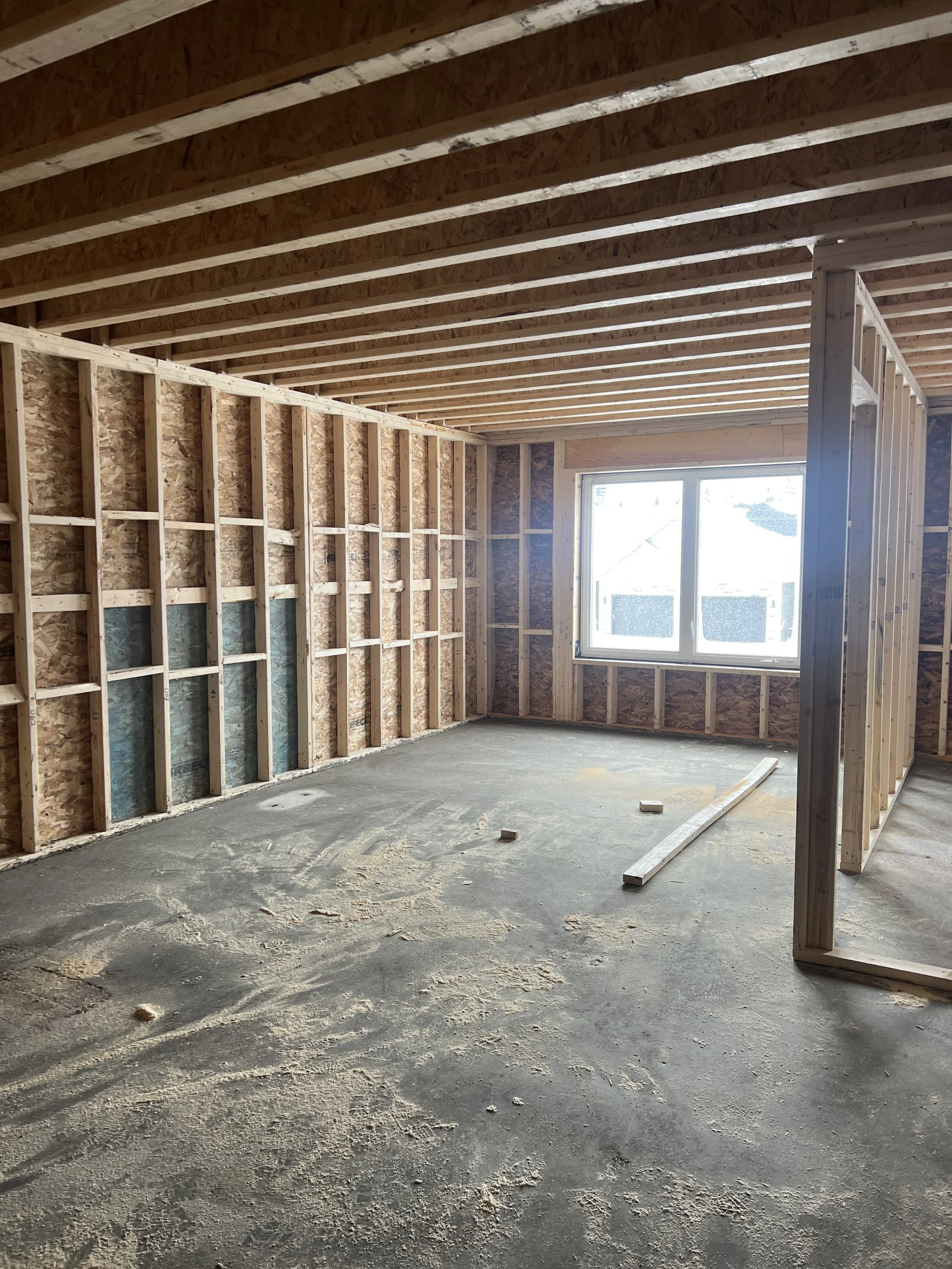 Interior view of a house under construction, with exposed wooden framing, a window, and a concrete floor.