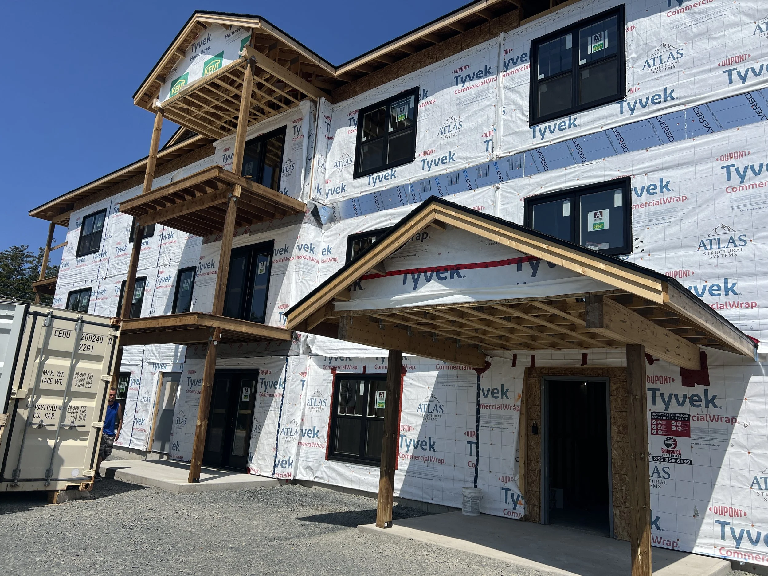 Construction site of a multi-story building with exterior walls covered in white Tyvek weather barrier wrap and black-framed windows, with wooden beams and a small front porch under construction.