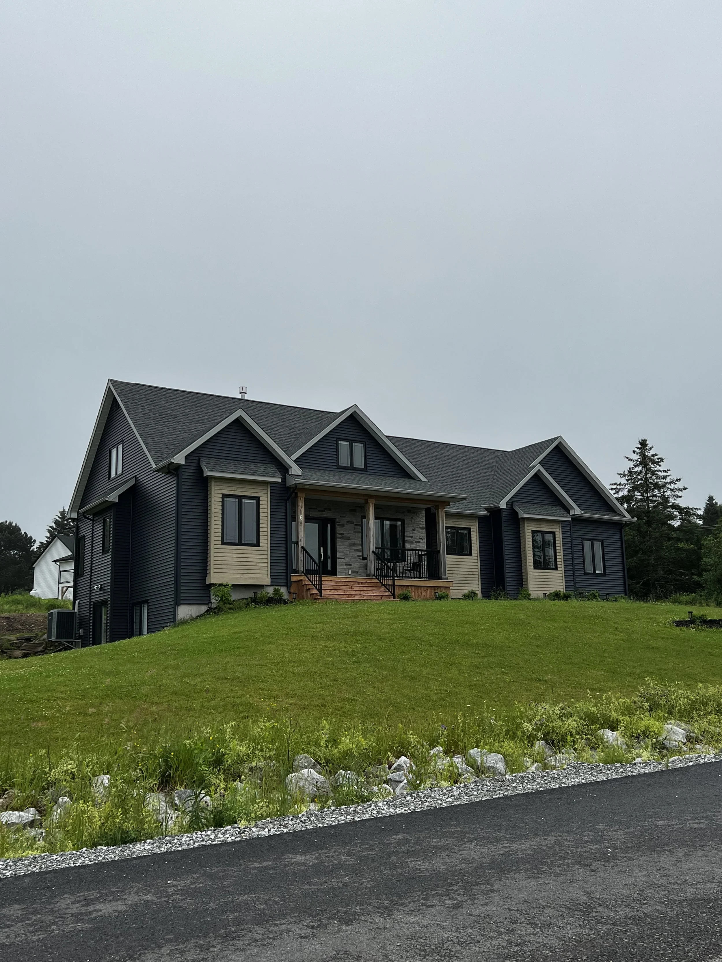 A modern two-story house with dark blue siding and beige accents, a covered front porch, and a gabled roof, situated on a green lawn with a gravel edge and a paved road in the foreground. There are trees in the background under an overcast sky.