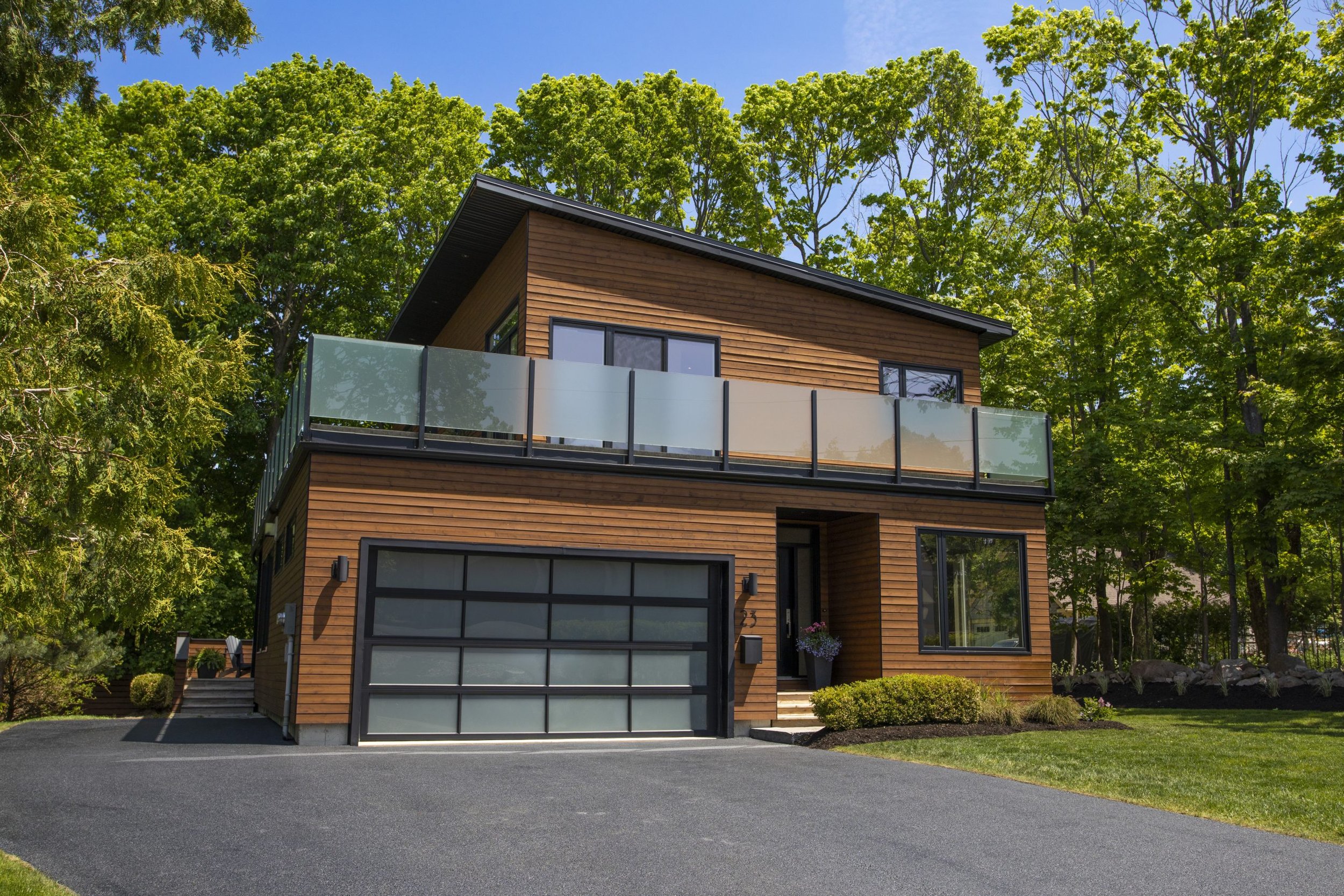 Modern wooden house with large black-framed windows and a balcony, surrounded by lush green trees, on a sunny day with a blue sky.