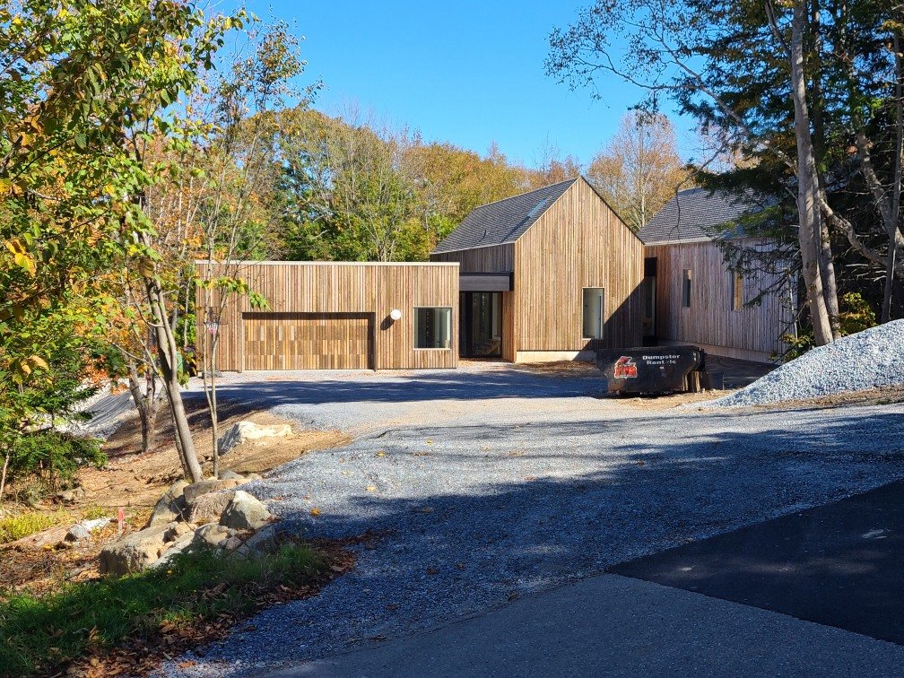 A modern wooden house under construction with a gravel driveway, surrounded by trees with fall foliage.