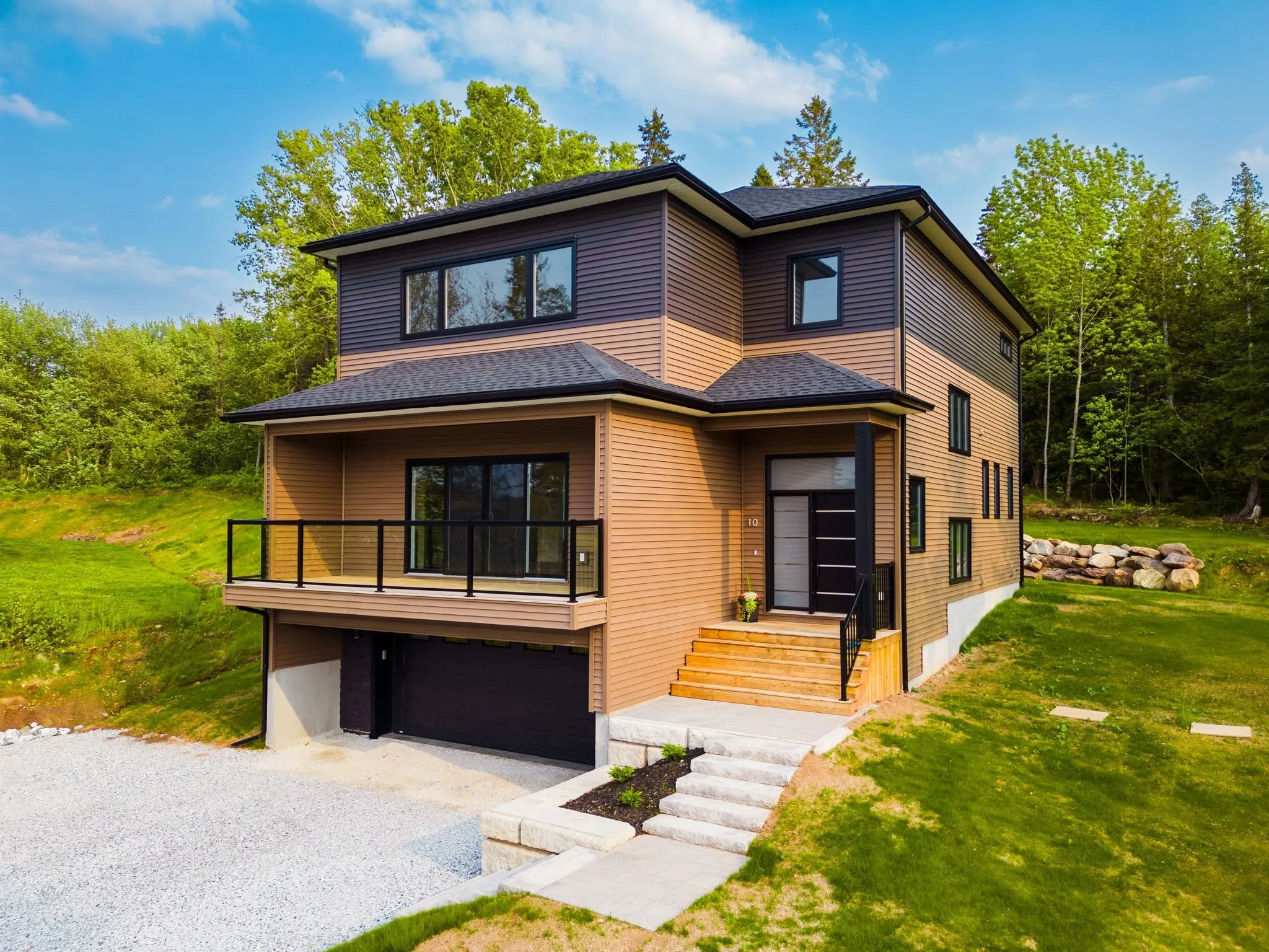 Modern two-story house with brown and black exterior, large windows, a garage, a small front porch with stairs, surrounded by green grass, trees, and a blue sky with clouds.