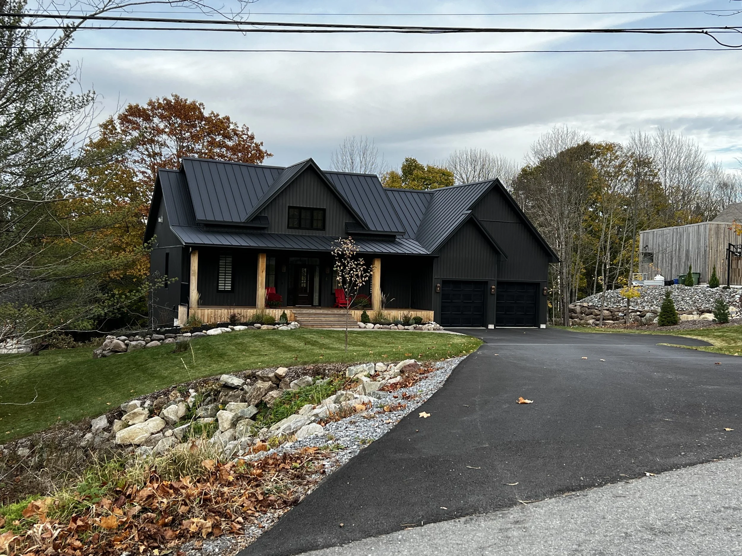 A modern black house with a metal roof and a front porch with red chairs, surrounded by trees with fall foliage, a fresh asphalt driveway, and landscaped rocks and plants.