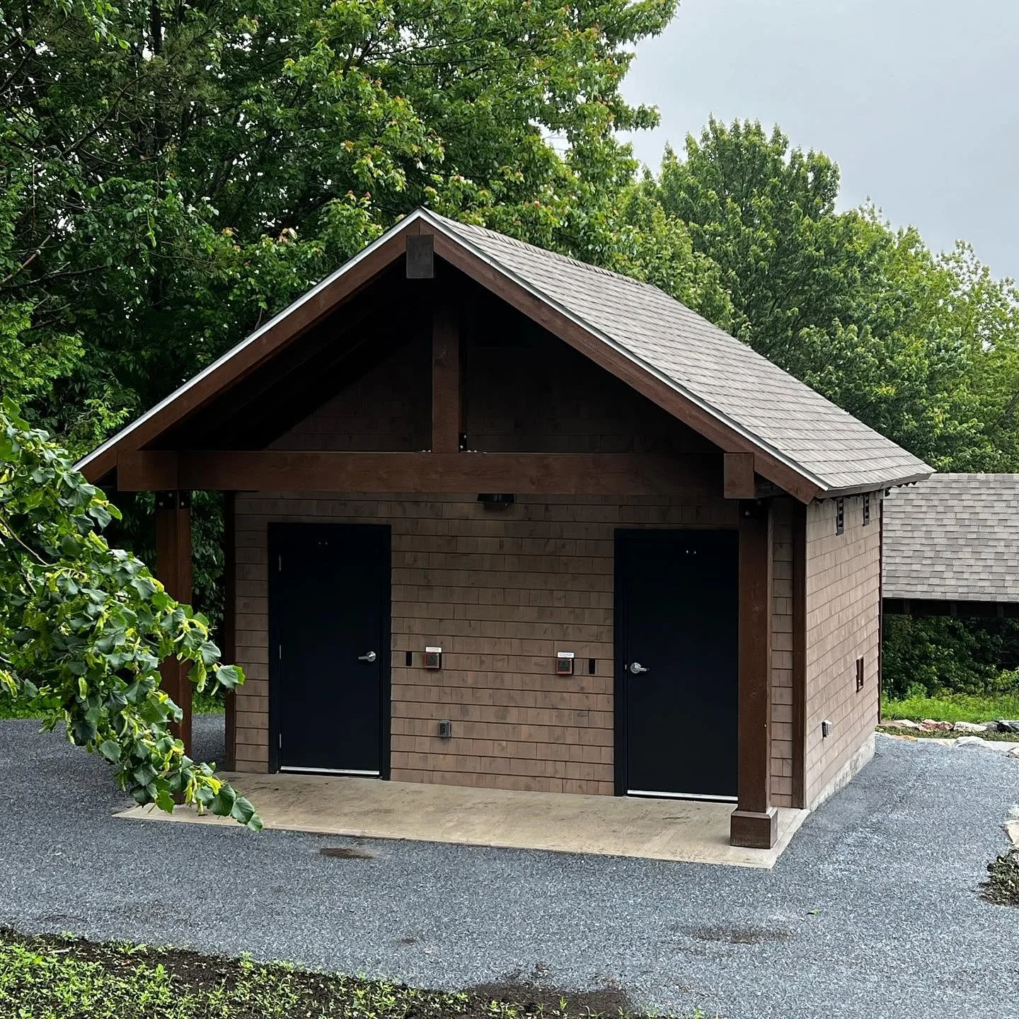 A small wooden building with a gabled roof, two black doors at the front, and surrounded by trees with green leaves.