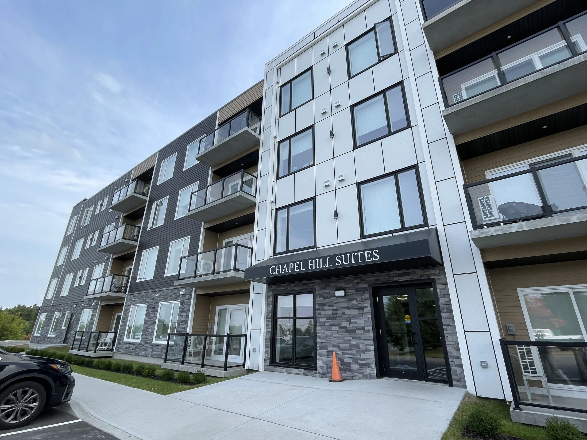 Modern multi-story apartment building with large glass windows and balconies, labeled 'Chapel Hill Suites,' with parking lot and a few parked cars in front.