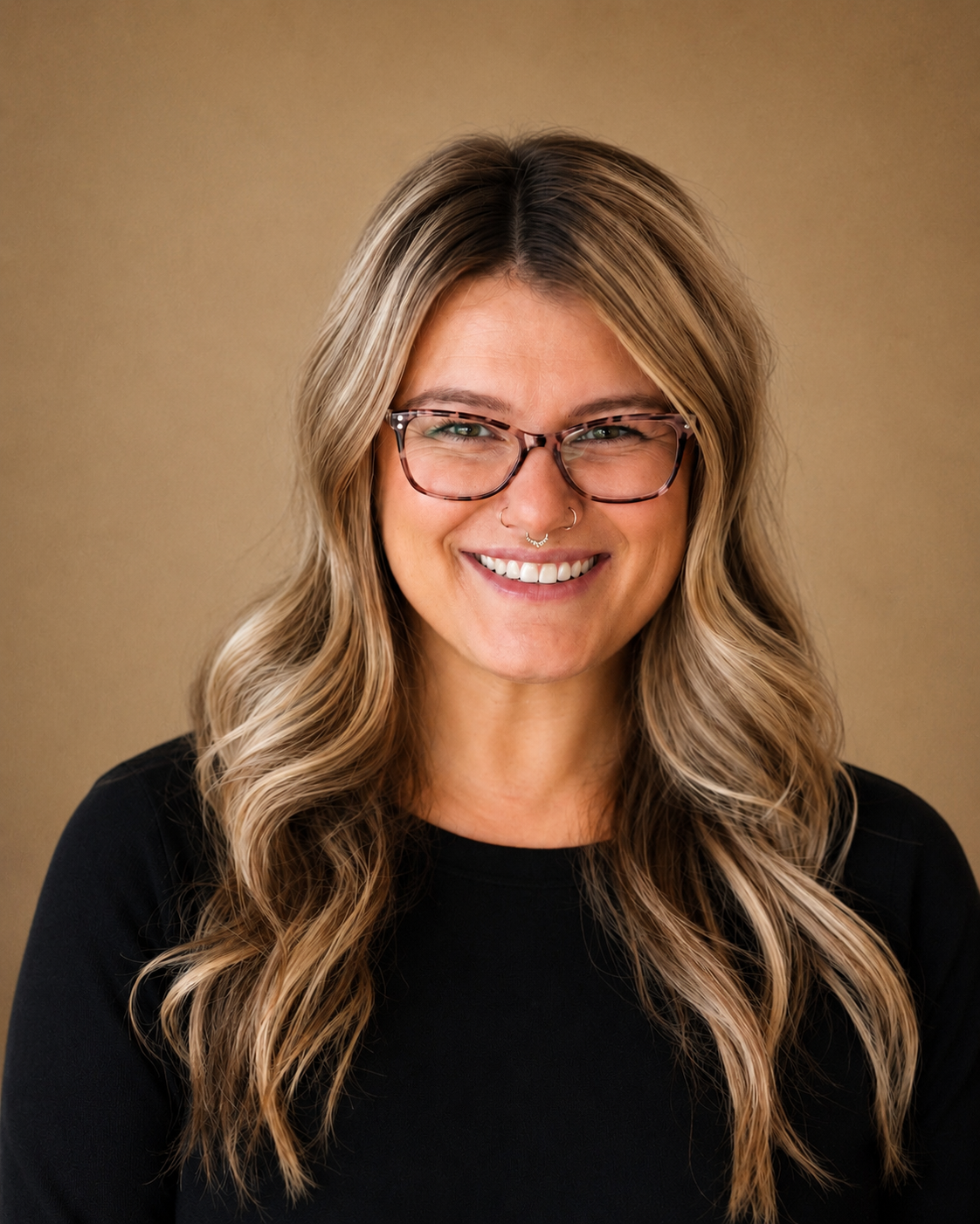 A woman with wavy, blonde hair, wearing glasses, smiling, against a tan background.