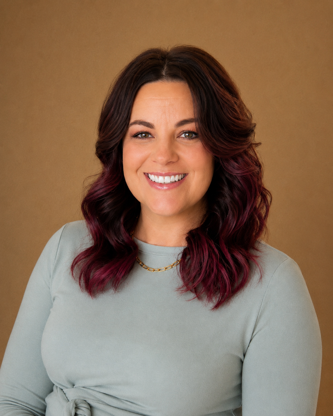 Smiling woman with brown and purple hair, wearing a light gray top and a gold chain necklace against a brown background.