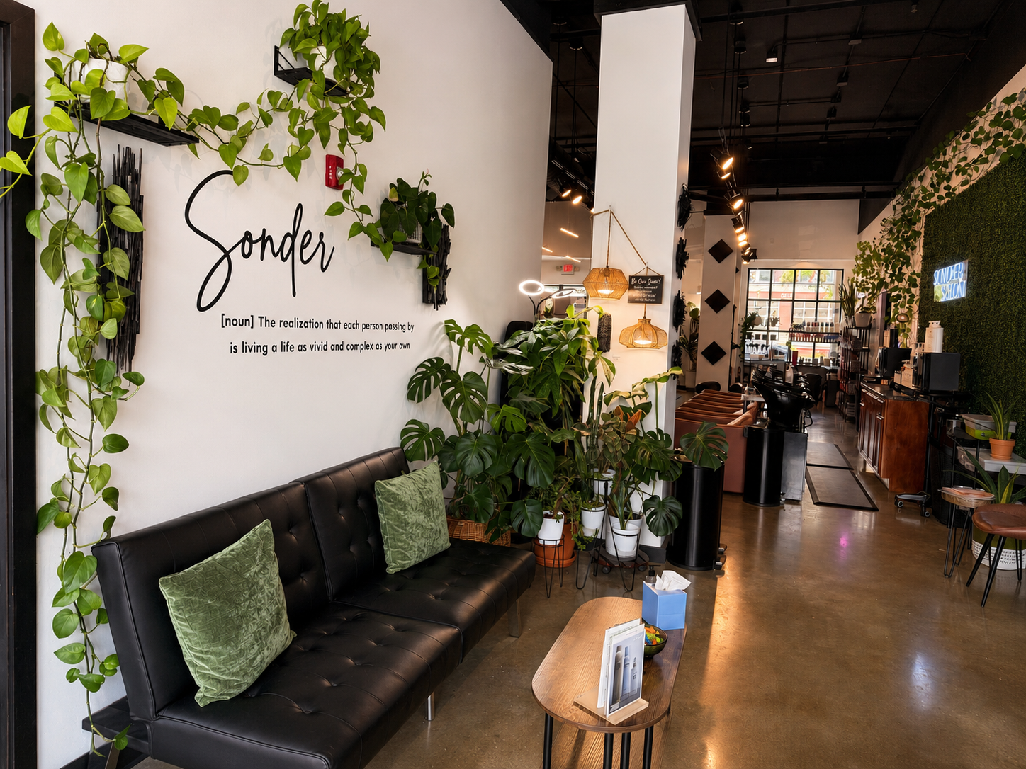 Interior of a modern cafe with a black leather couch, green pillows, numerous houseplants, and decorative hanging lights. There's a wall with the word 'Sonder' and a definition, with seating and workspace visible in the background.