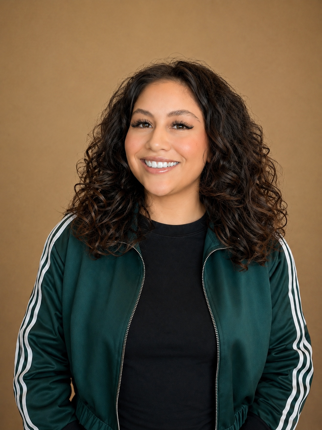 A young woman with long, curly dark hair smiling while wearing a black shirt and a green zip-up jacket with white stripes on the sleeves, standing against a plain brown background.