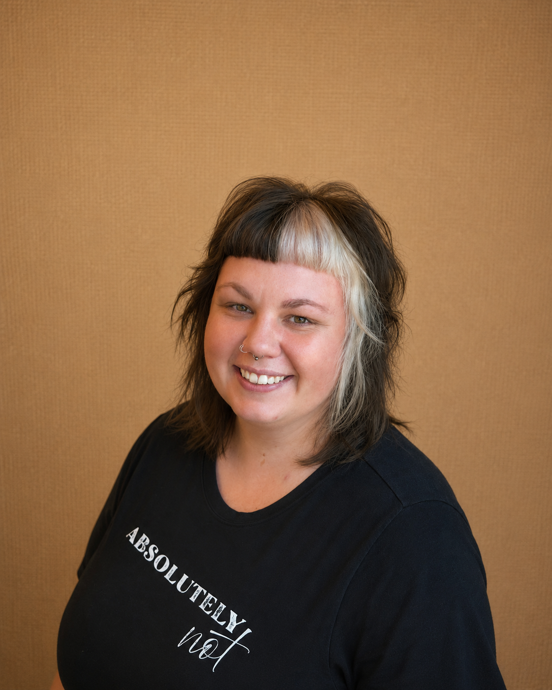 A smiling woman with light skin, dark and blonde hair, and a septum piercing, wearing a black T-shirt with white text, standing against a brown background.