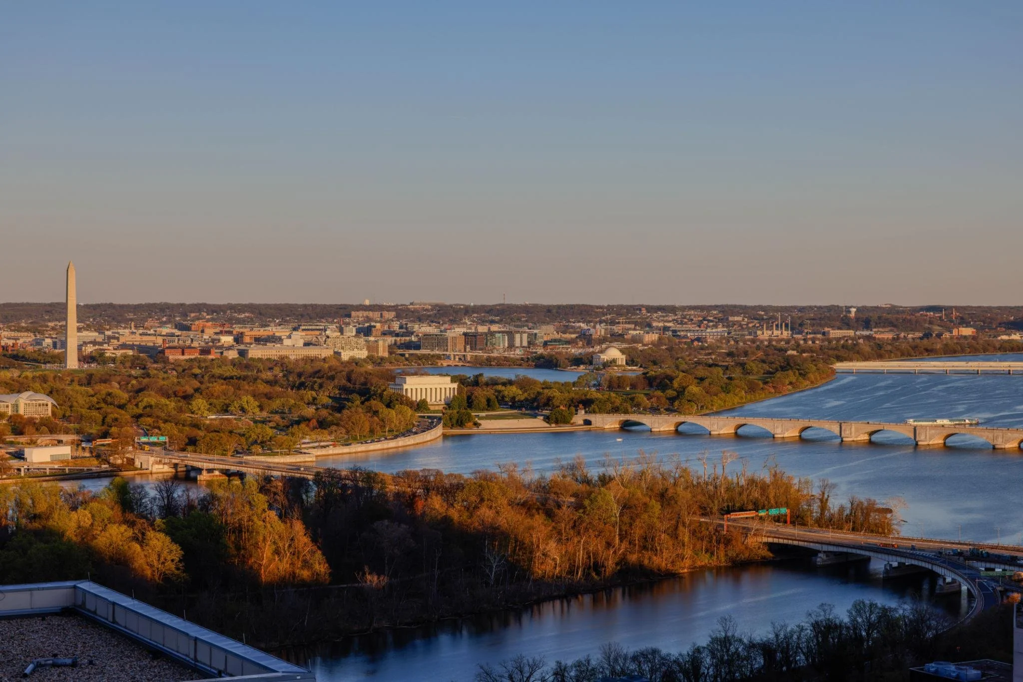 View of Washington, D.C. skyline during sunset with the Washington Monument visible on the left, the Potomac River in the foreground, and trees with fall foliage.