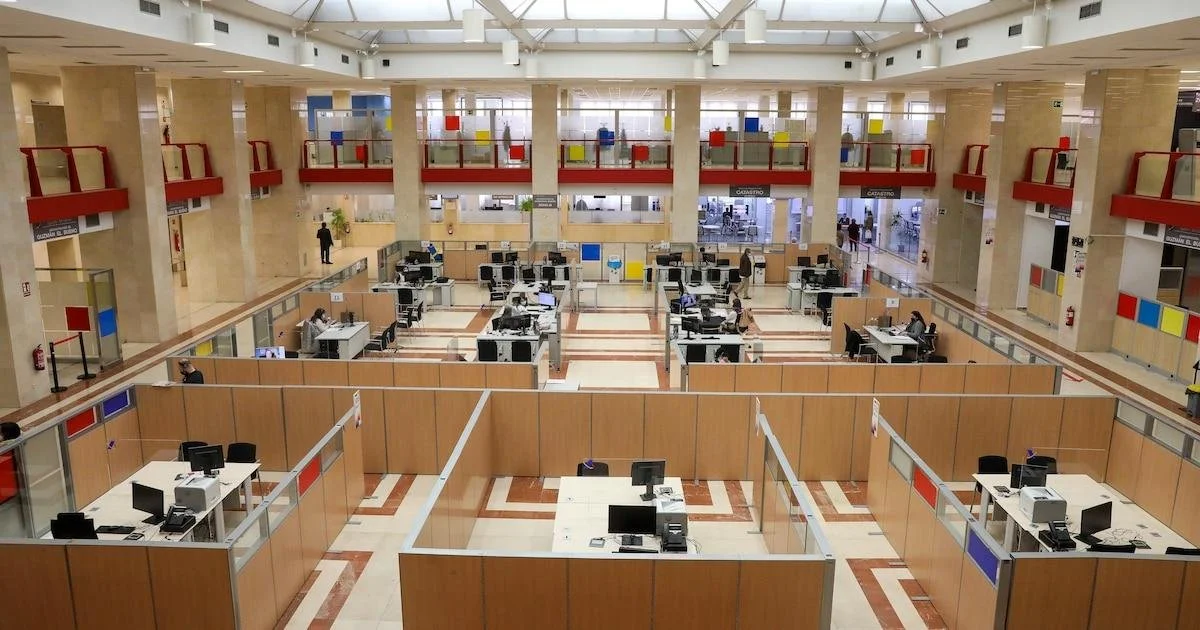 Empty cashier booths with computers inside a large, spacious billing or service area of an airport or government office, with a few employees and visitors.
