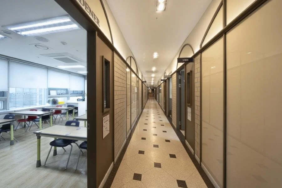 A school hallway with a classroom visible on the left side, featuring desks and chairs near large windows. The corridor has a patterned linoleum floor and beige walls with black framing, leading to a vanishing point in the distance.
