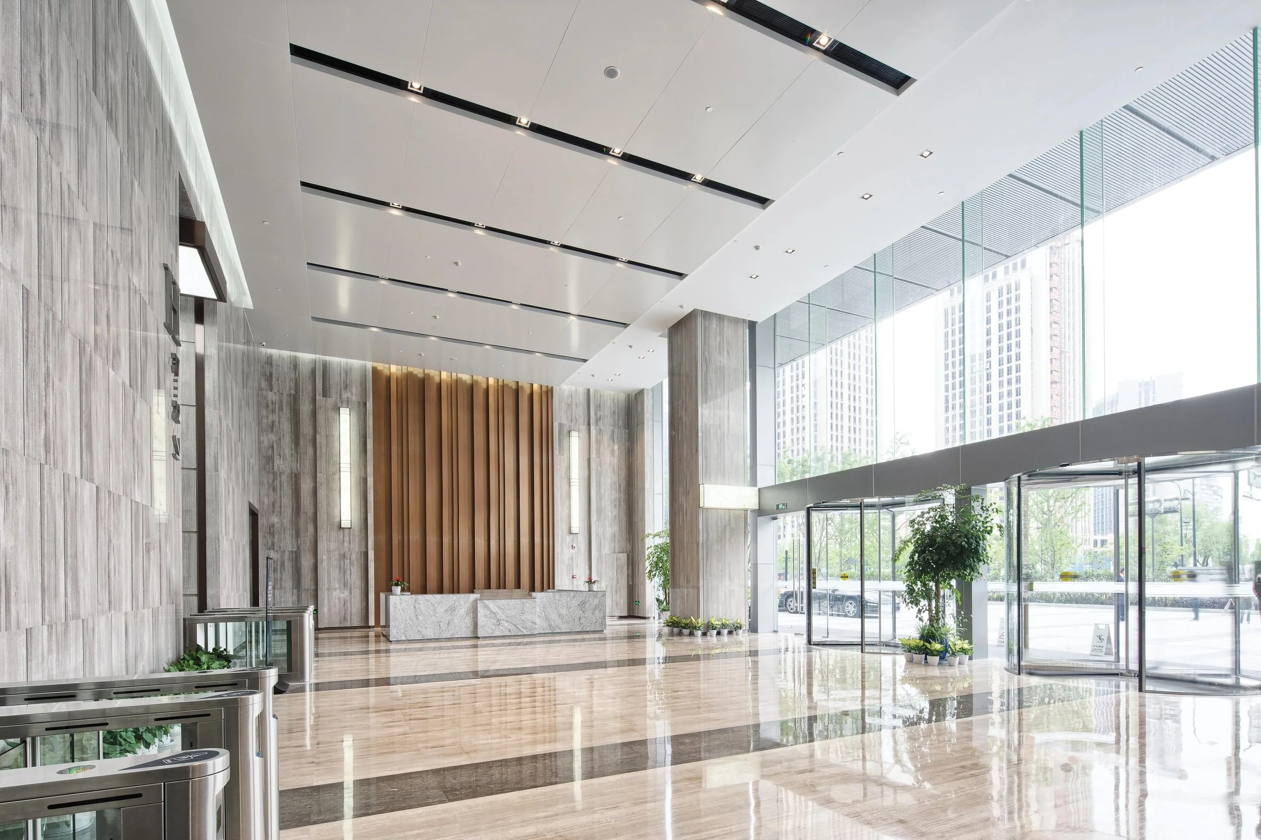 Modern lobby with high ceilings, large floor-to-ceiling windows, marble floors, and minimalistic decor. There are a few potted plants, a marble reception desk, and some wall lighting accents.