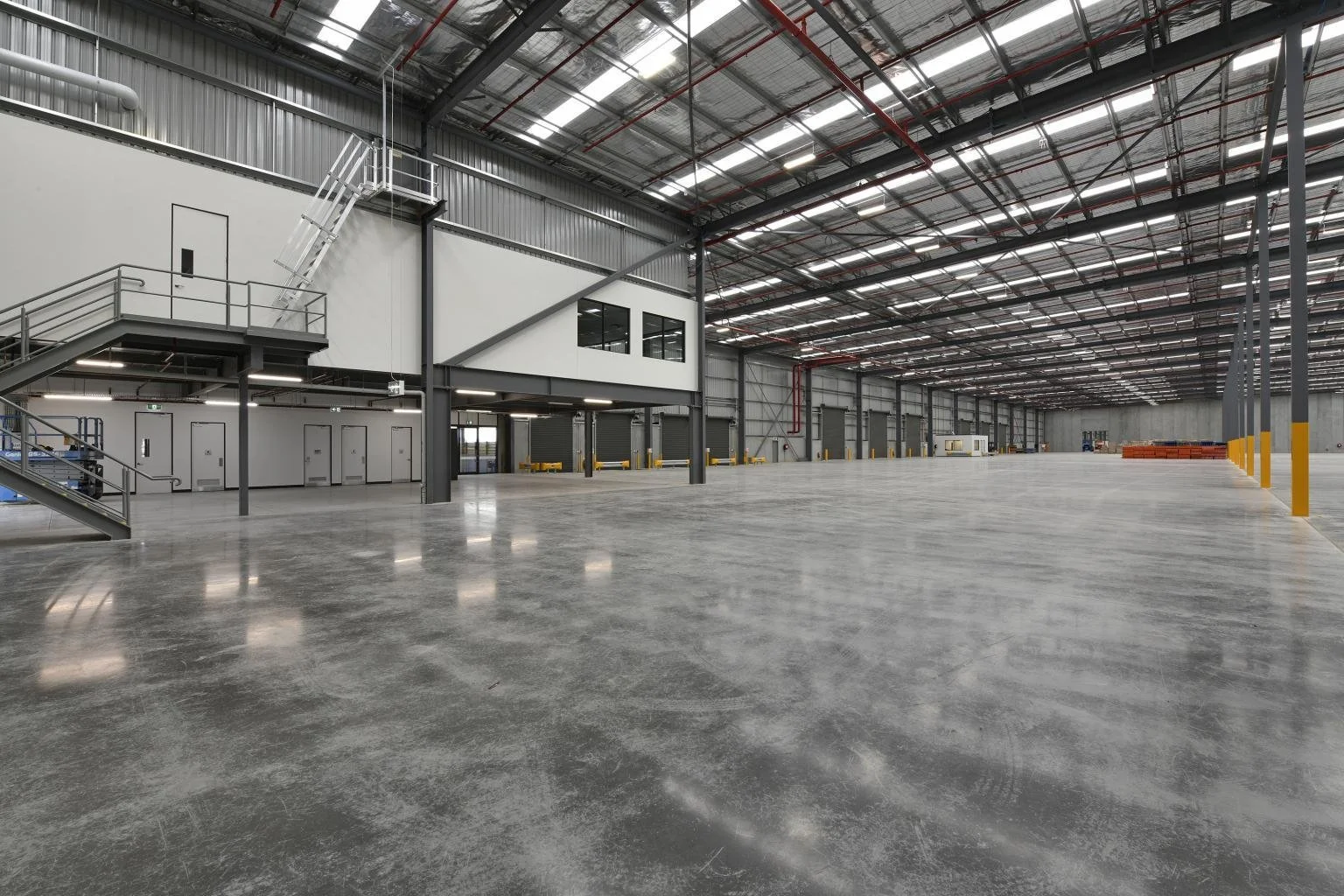 Interior of a large, empty warehouse with polished concrete floors, high ceilings with metal beams, and unfinished walls. There are some office spaces and a staircase on one side, and a few scattered items along the walls.