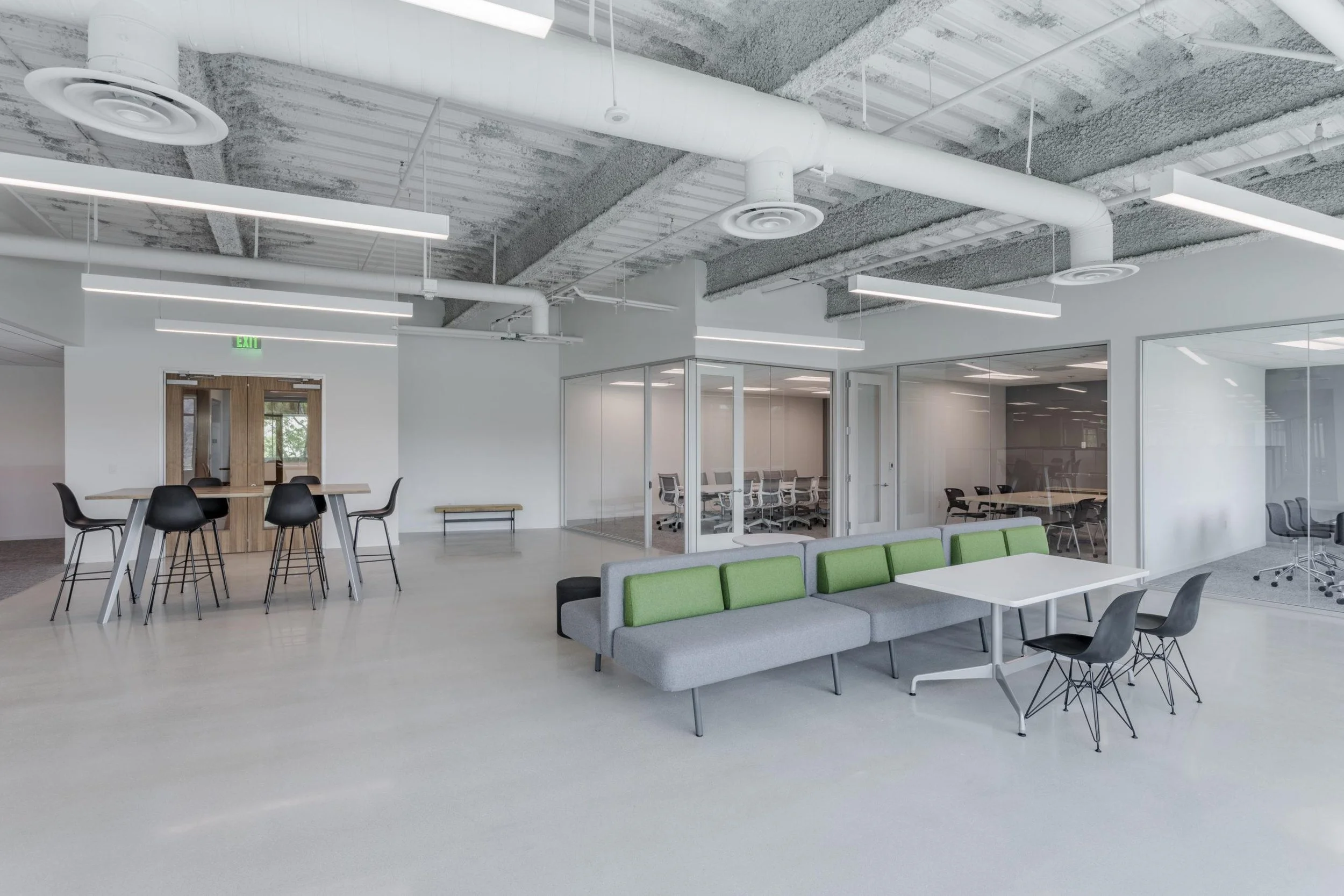 Modern, open-plan office space with exposed ceiling pipes and industrial lighting, featuring conference rooms with glass walls, lounge seating, and meeting tables.